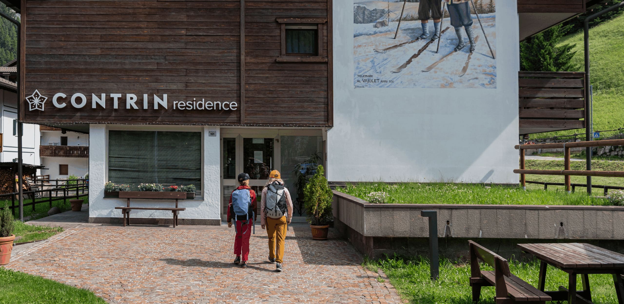 Edificio di Contrin Residence, due escursionisti con zaini entrano dalla porta
