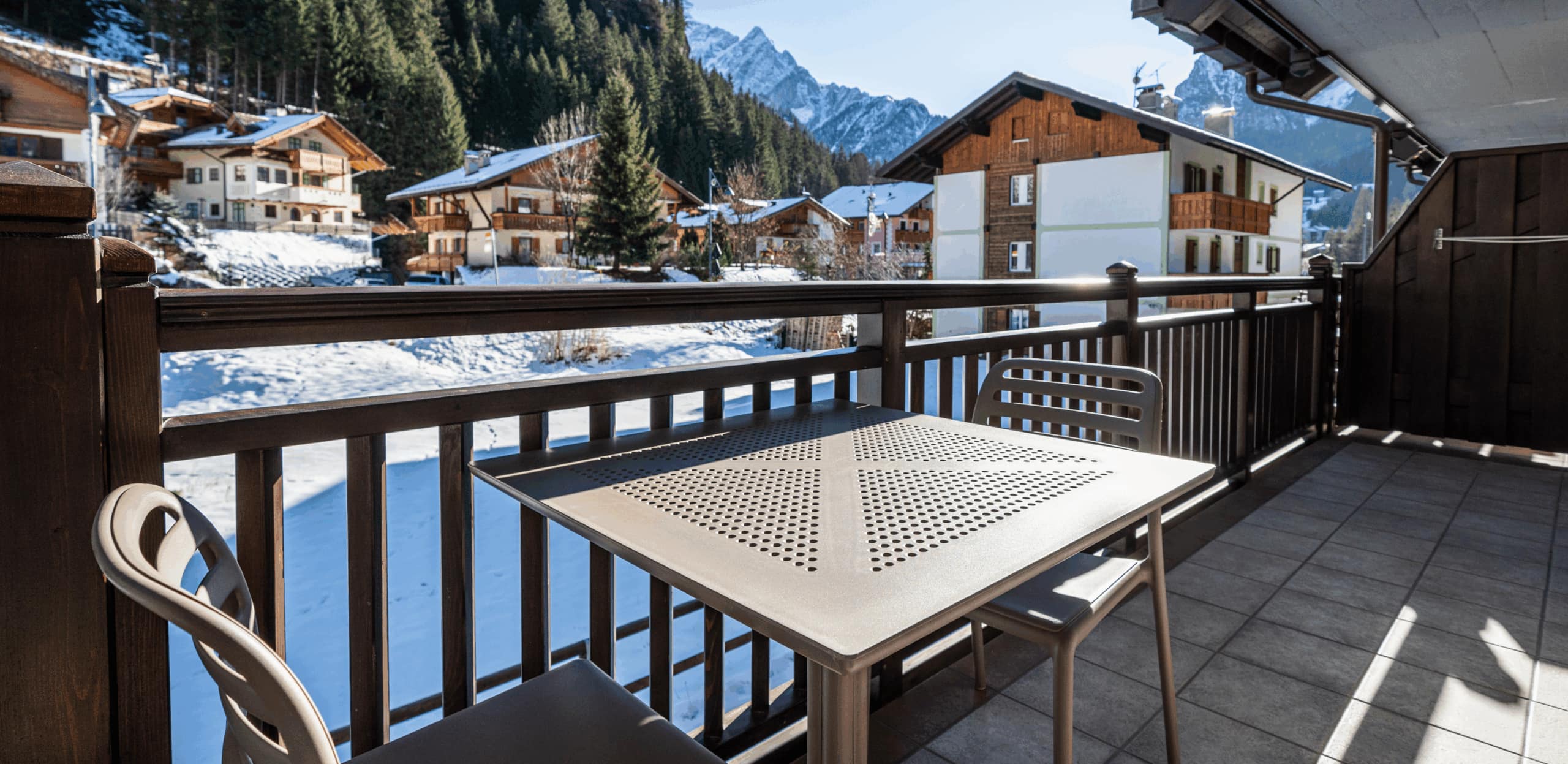 Balcone con tavolo e sedie metalliche che guarda su case alpine innevate e montagne sullo sfondo