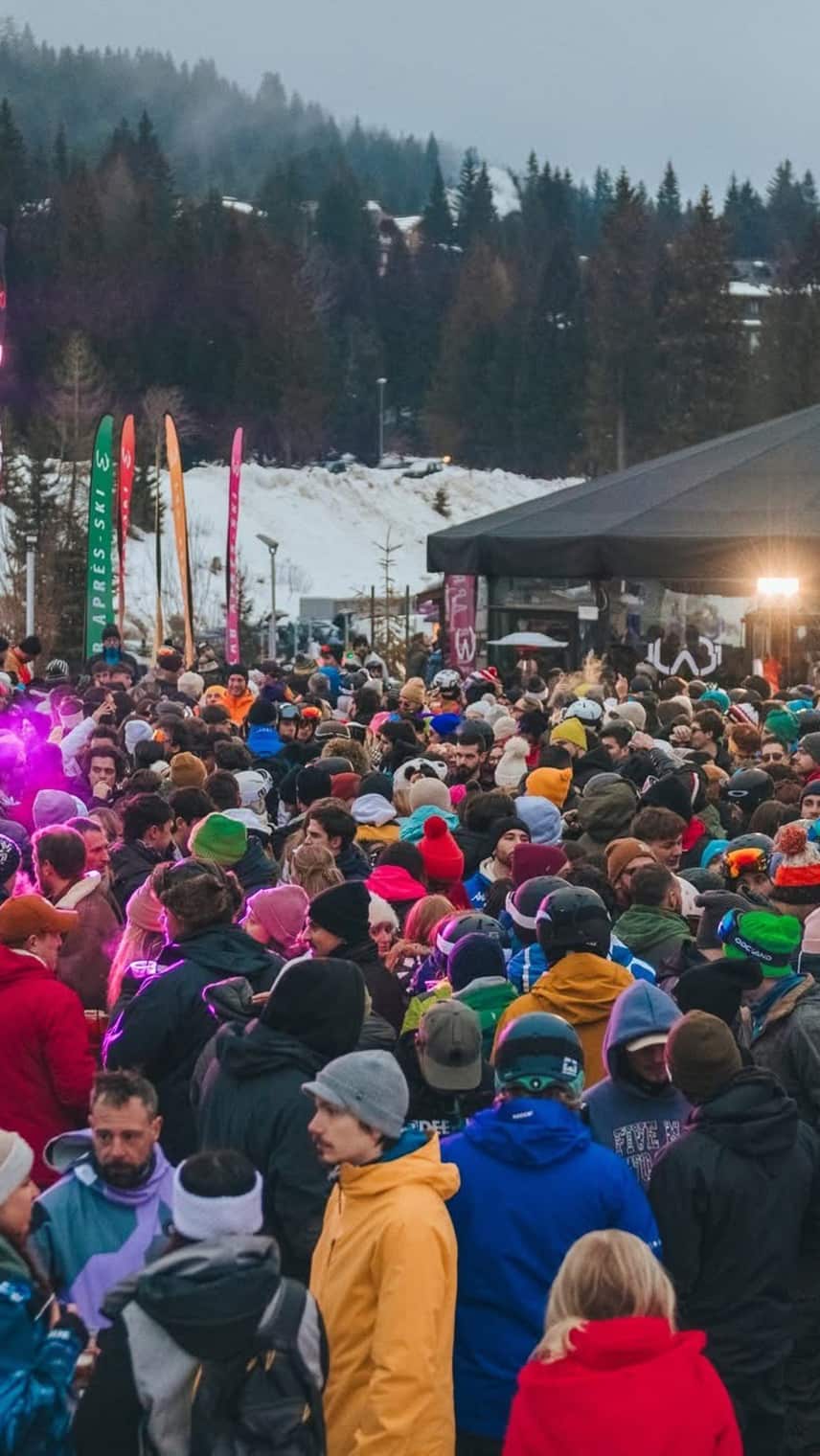 Menschenmenge bei einer Außen‑Après‑Ski‑Party vor Bühnenzelt; verschneite Hänge und Tannen im Hintergrund.