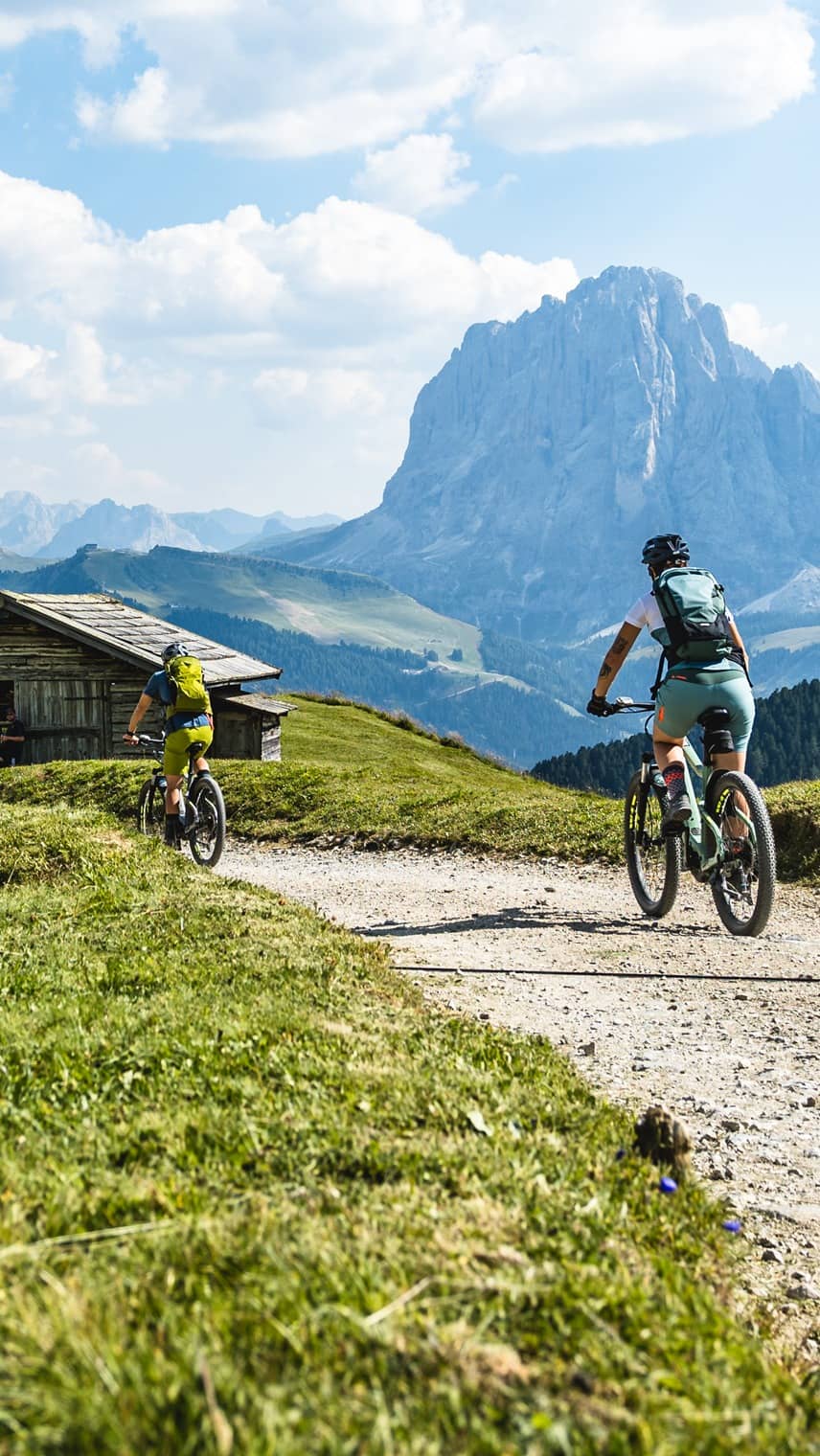 Zwei Mountainbiker auf Schotterweg in alpiner Wiese, Holzhütte links, massiver Berg im Hintergrund.