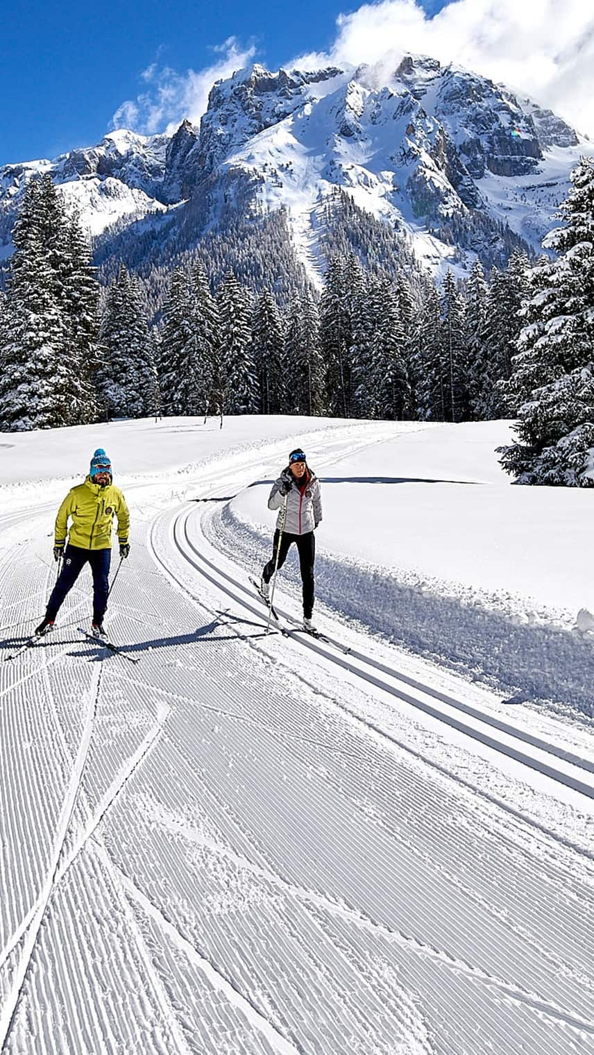 Zwei Langläufer auf präparierter Spur vor schneebedeckten Tannen und hoch aufragendem Berg