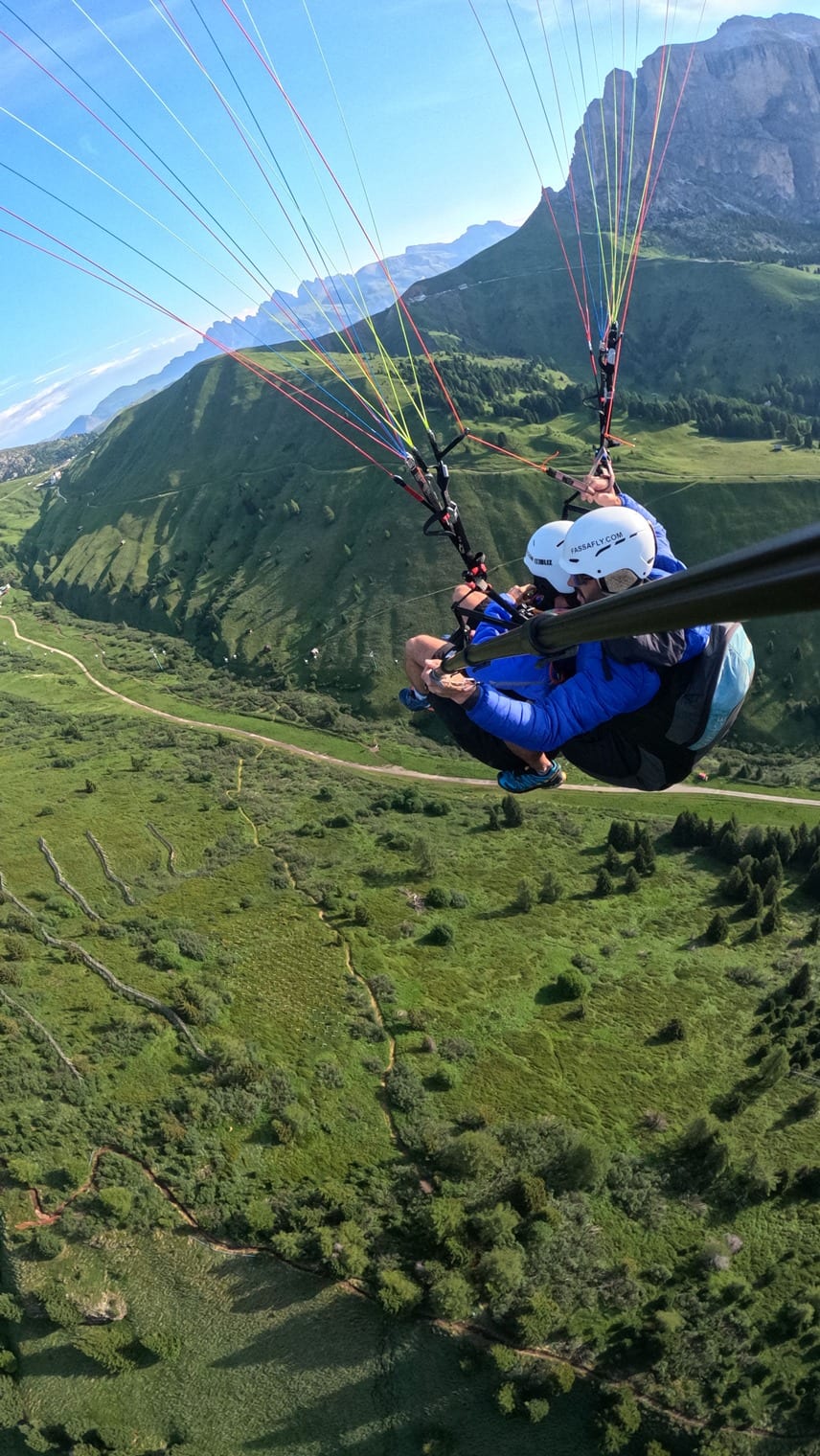 Tandem-Gleitschirmflug über grünem Bergtal, zwei Personen mit Helmen, bunte Leinen, Bergkamm im Hintergrund