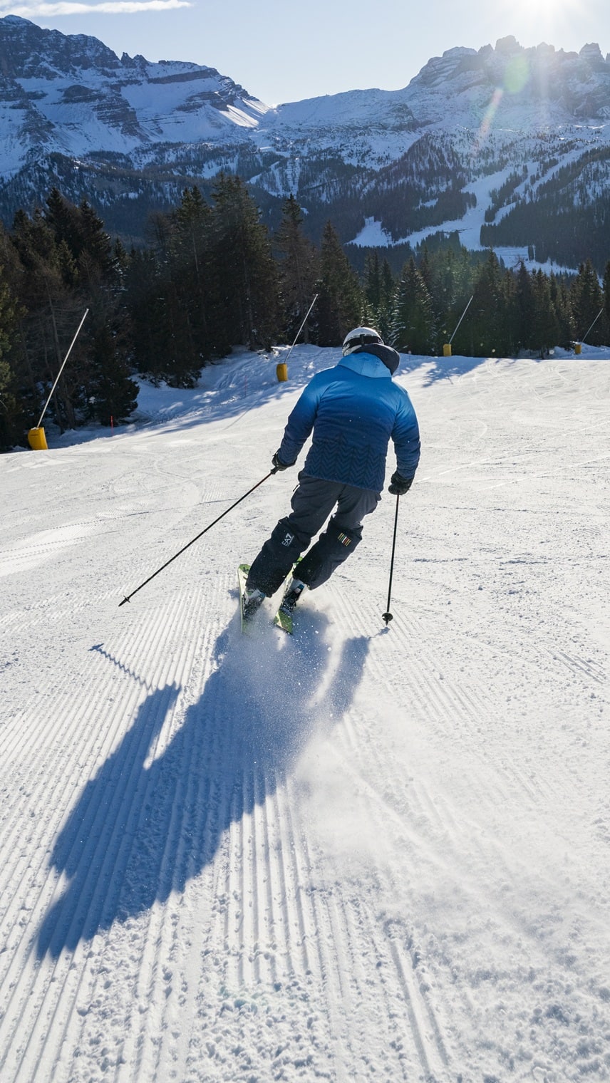 Skifahrer in blauem Anorak fährt eine präparierte Piste bergab; verschneite Tannen und Berggipfel im Hintergrund