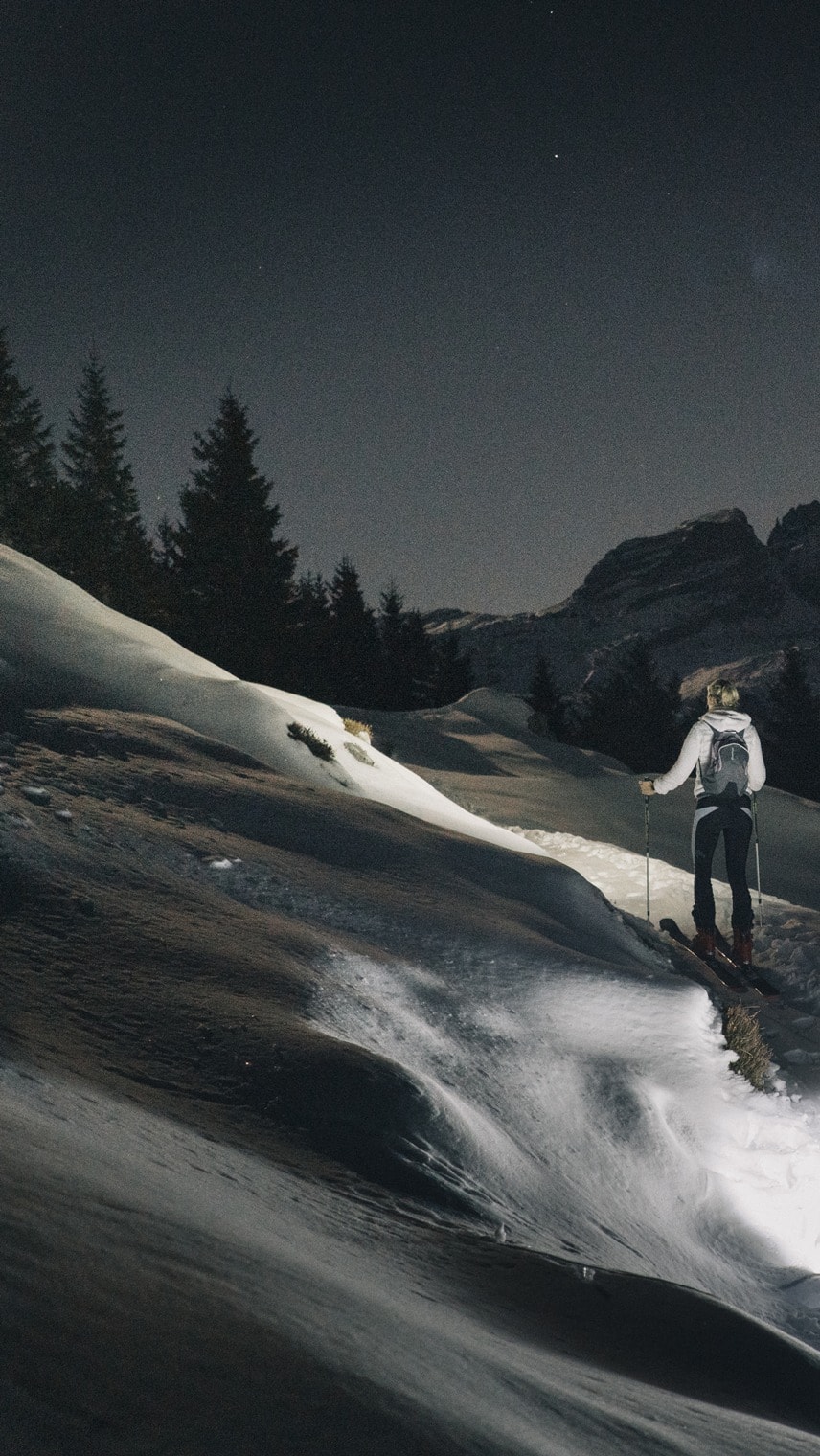 Skitourengeher steigt nachts einen verschneiten Hang hoch, Licht beleuchtet Spur, Tannen und Bergsilhouette