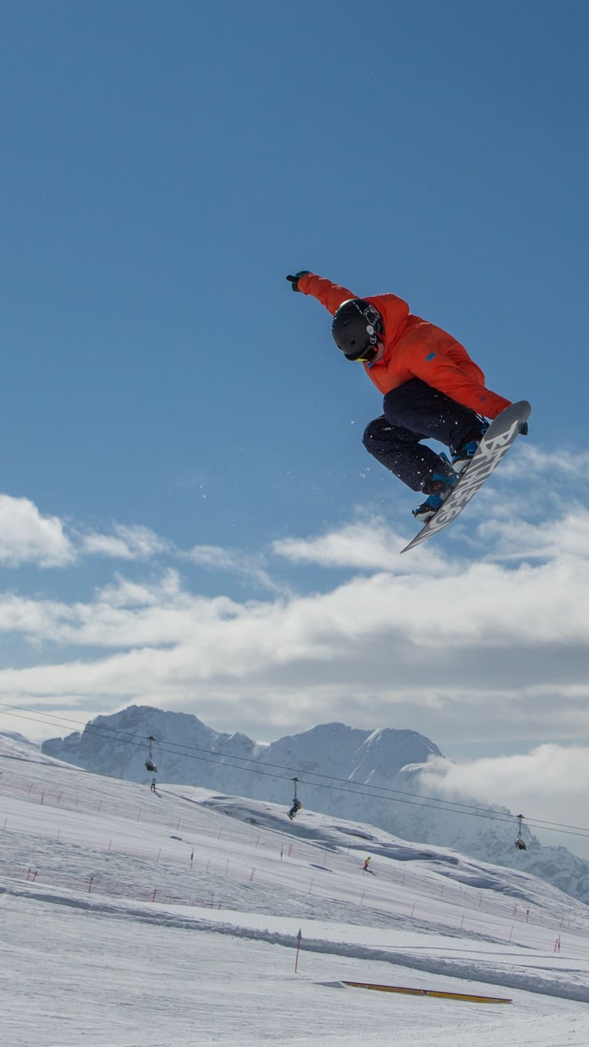 Snowboarder in orange Jacke mit Helm beim Sprung über verschneite Piste, Berge und Sessellift im Hintergrund.