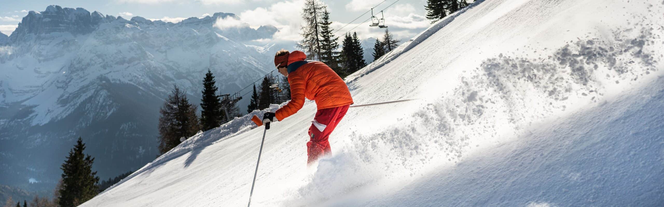 Sciatore in giacca arancione scende su ripida neve polverosa con montagna innevata e seggiovia sullo sfondo