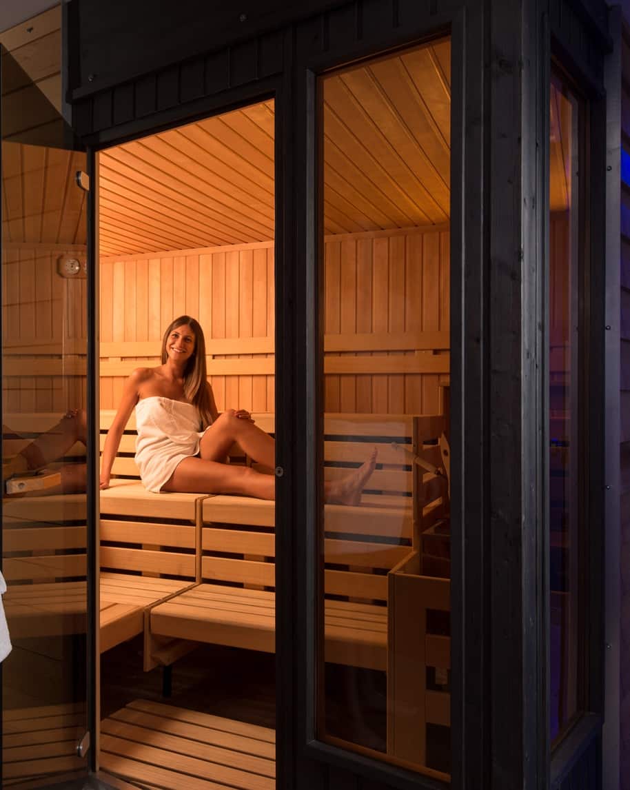 Donna in asciugamano seduta su panca in sauna di legno, vista attraverso porta vetrata