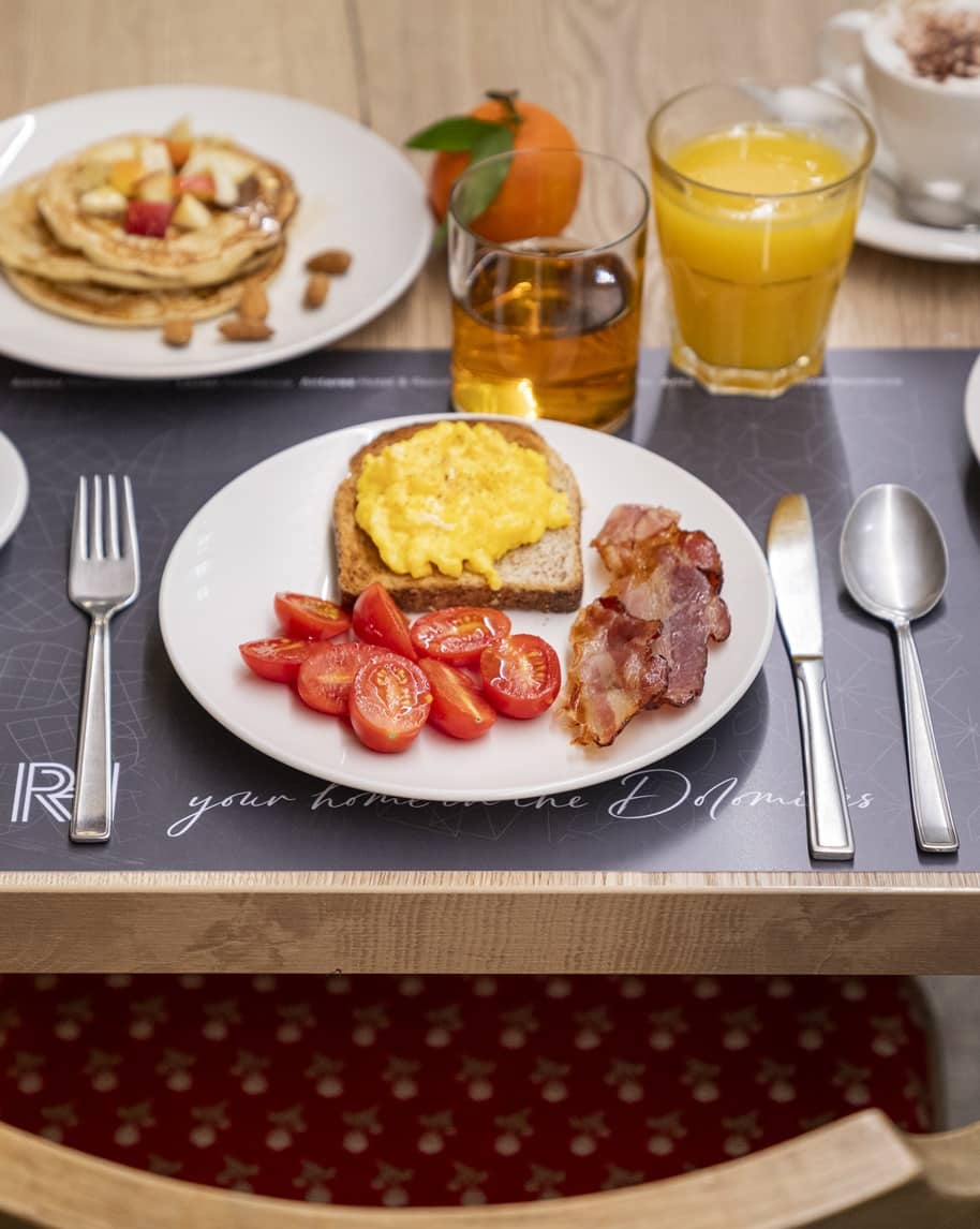 Toast con uova strapazzate, pomodorini e bacon su piatto; sul tavolo bicchiere di succo d'arancia e pancake