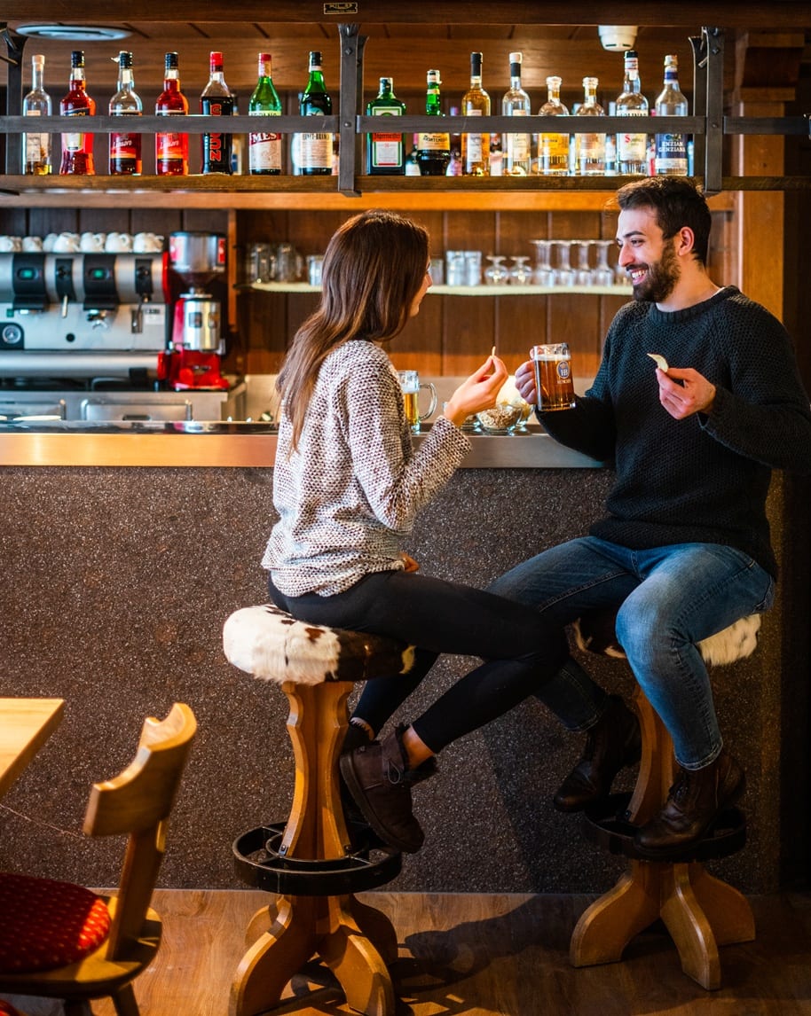 Coppia al bancone del bar, sorridono bevendo birra e mangiando snack; bottiglie su mensola dietro