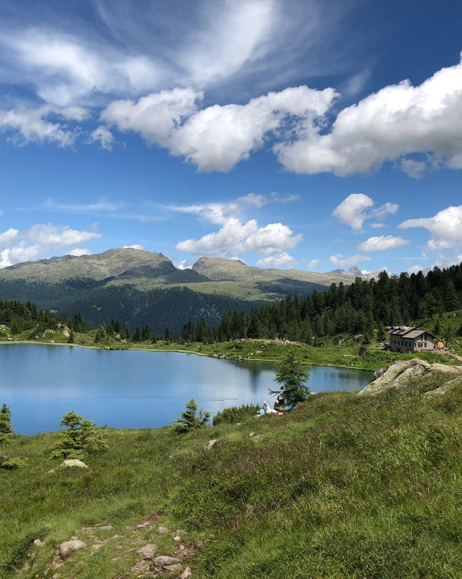 Lago alpino circondato da prati e pini, baita sulla riva e montagne sullo sfondo sotto cielo nuvoloso