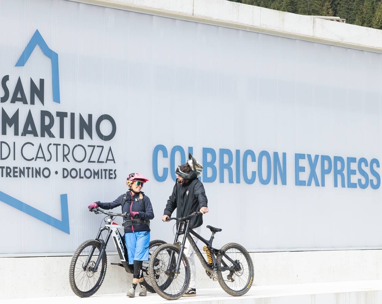 Due ciclisti con mountain bike in piedi davanti alla scritta Colbricon Express a San Martino di Castrozza
