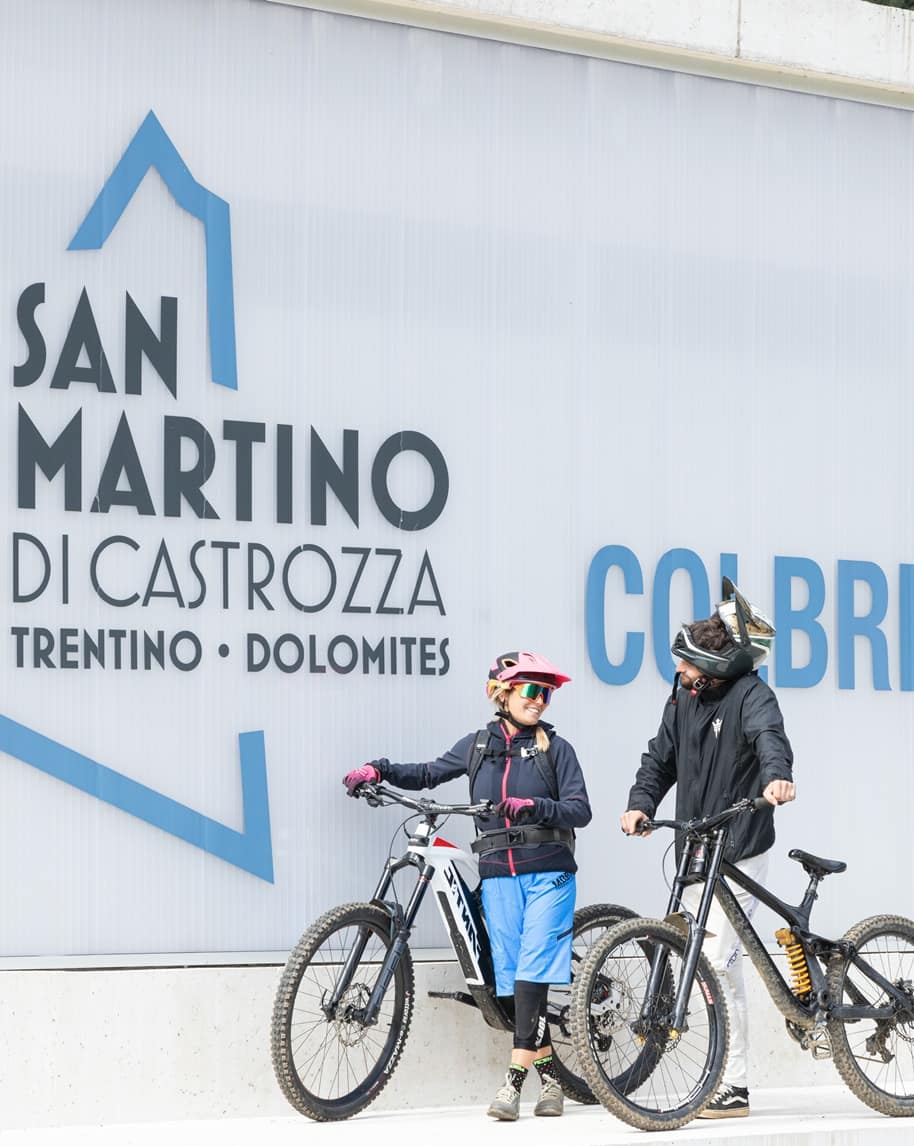 Due ciclisti appoggiati alle e-bike davanti al cartello SAN MARTINO DI CASTROZZA