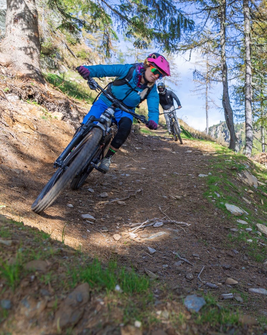 Due ciclisti in discesa su sentiero boschivo, primo con casco rosa in curva, secondo a seguire