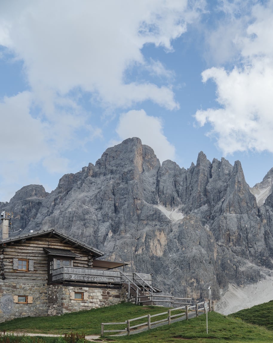 Baita in legno su prato con ringhiera davanti a parete rocciosa delle Dolomiti sotto cielo nuvoloso