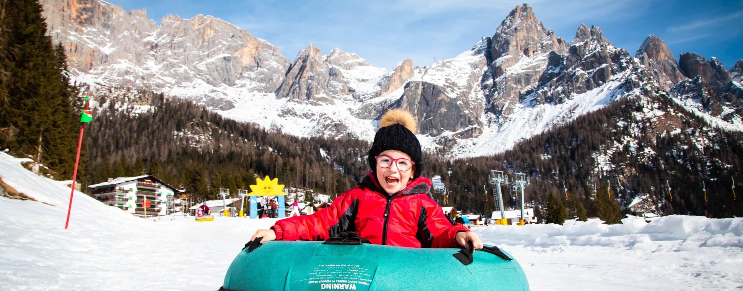 Bambino su ciambella da neve sorridente con giacca rossa, montagne innevate e impianti di risalita sullo sfondo