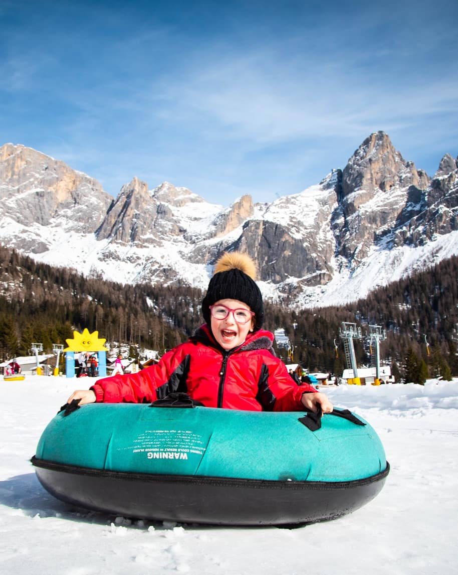 Bambino su ciambella da neve sorridente con giacca rossa su pista, montagne innevate sullo sfondo