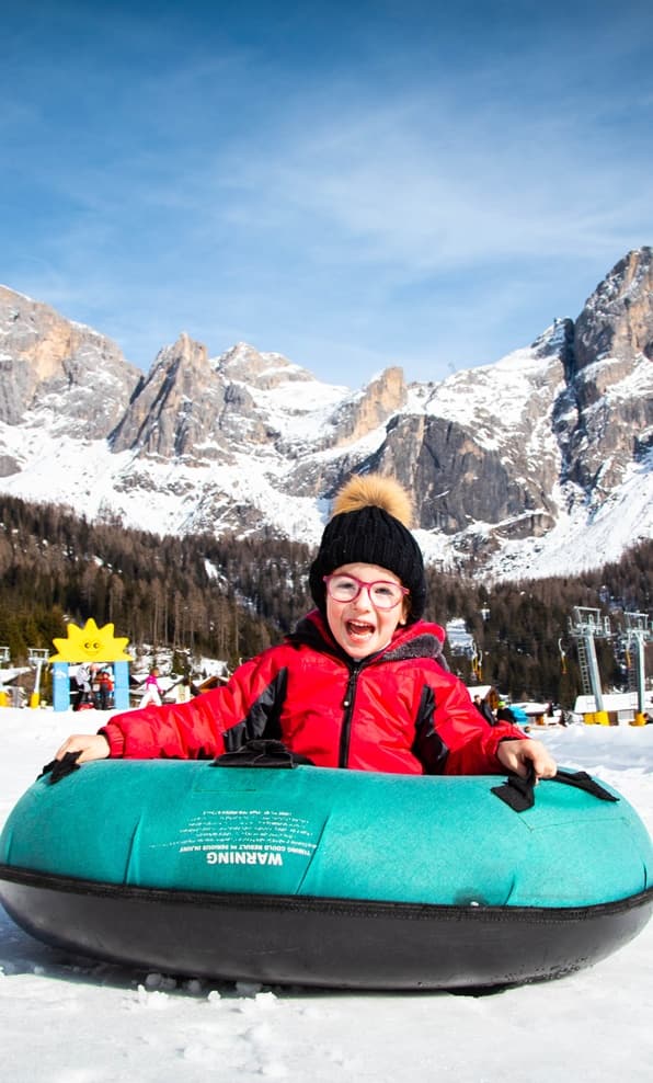 Bambino con giacca rossa e berretto nero su ciambella da neve turchese, su pista innevata con montagne sullo sfondo