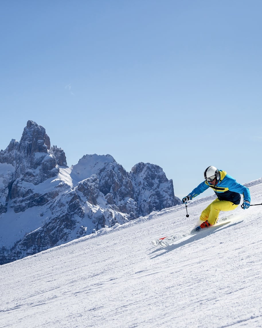 Sciatore in giacca blu e pantaloni gialli che curva su pista innevata, montagne rocciose innevate sullo sfondo
