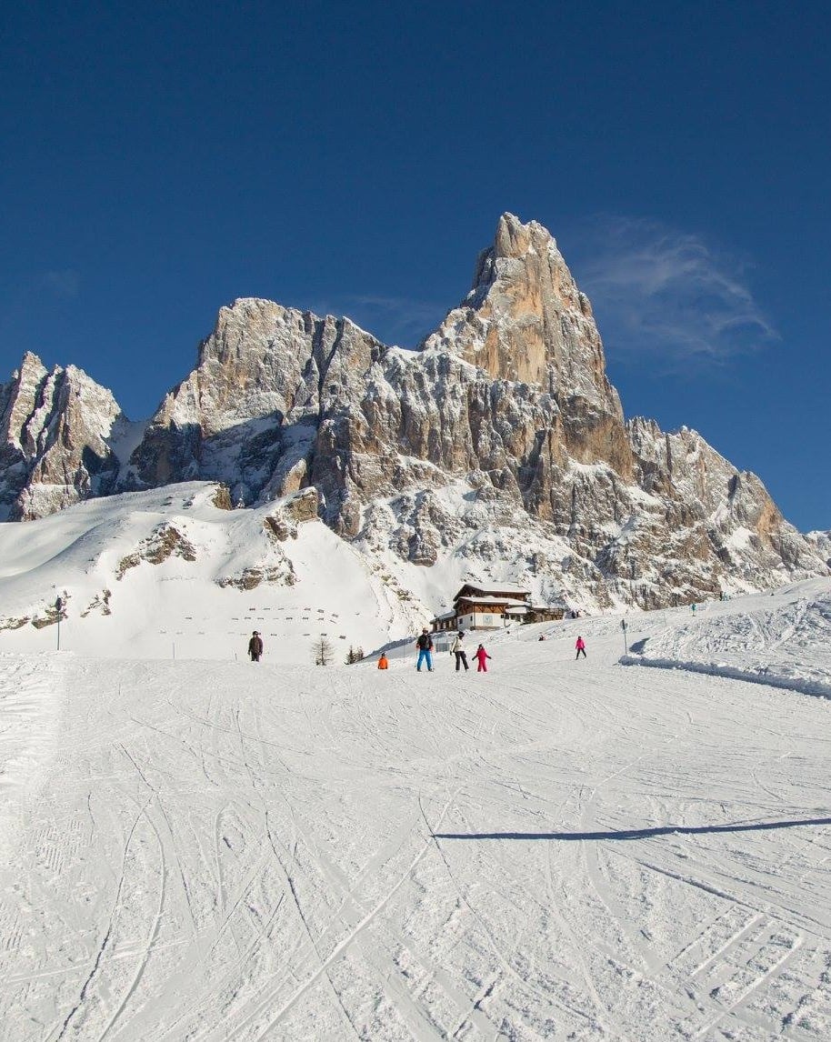 Cima rocciosa innevata, sciatori su pista con chalet ai piedi e cielo blu.
