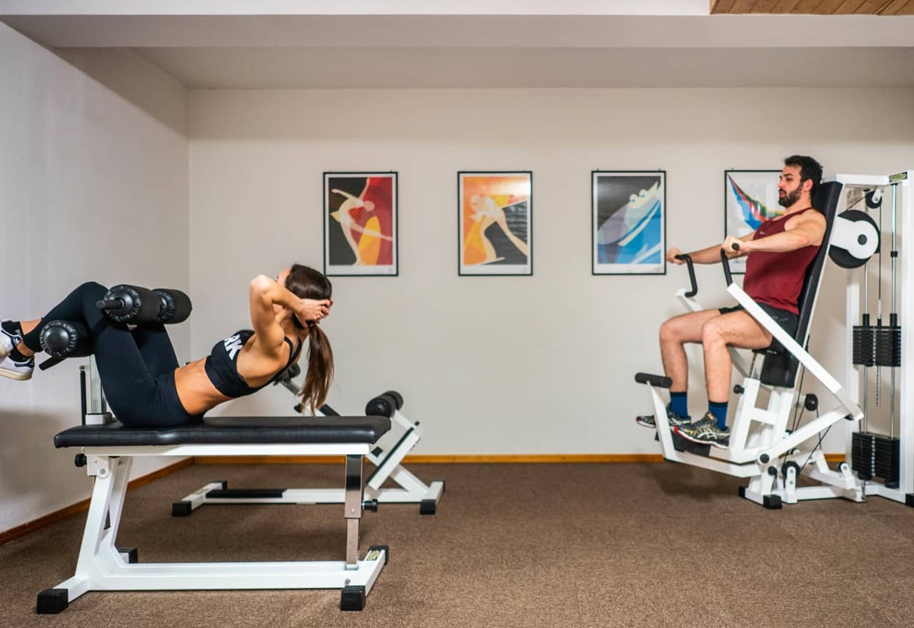 Donna su panca esegue addominali; uomo a destra usa macchina per petto in palestra