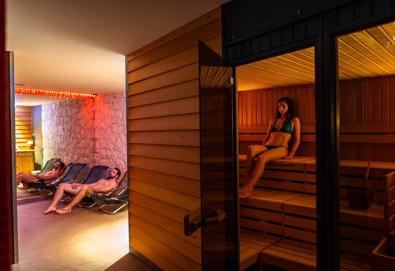 Donna seduta su panca di legno in sauna, porta in vetro aperta; a sinistra due uomini sdraiati su lettini relax