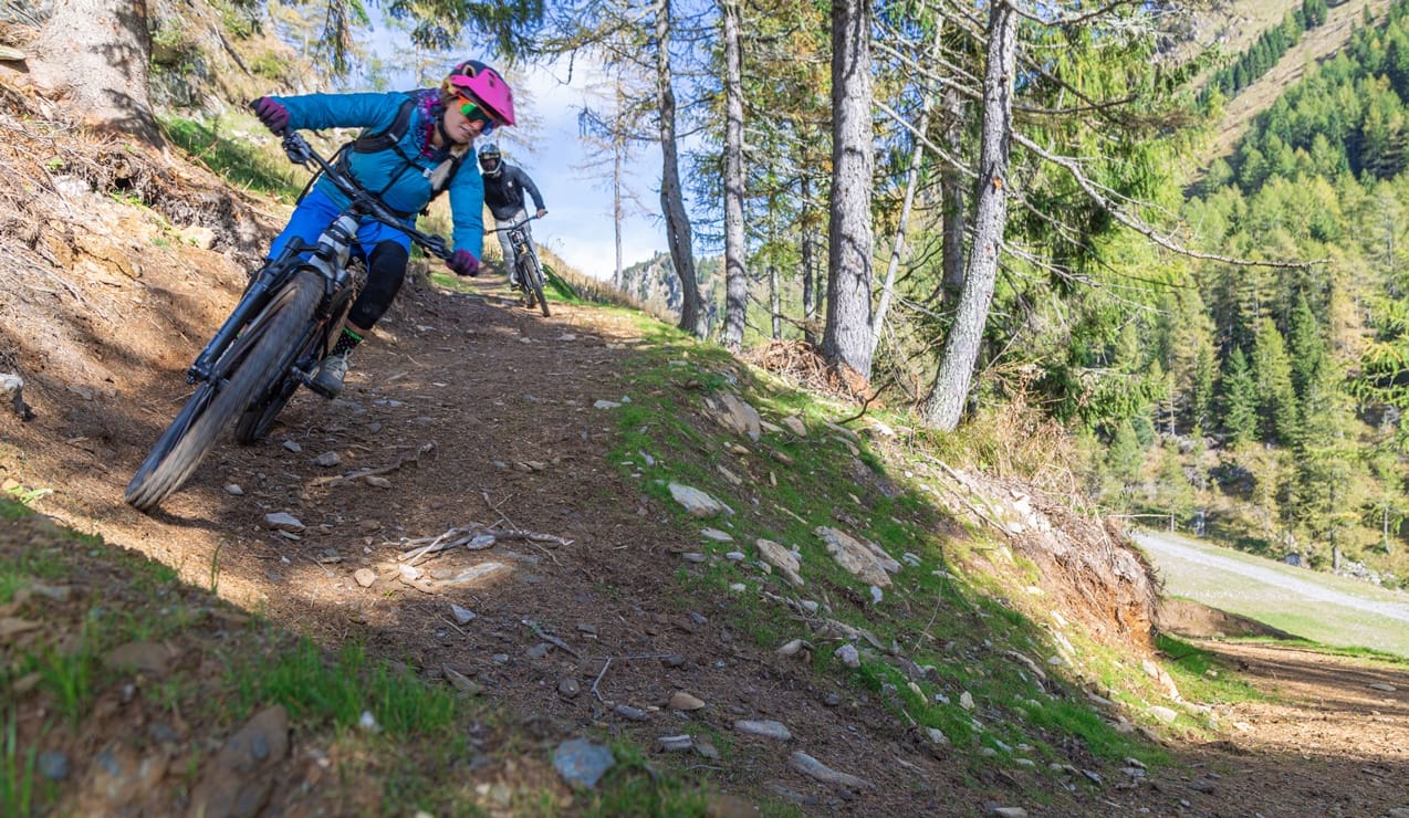 Due ciclisti in mountain bike che affrontano una curva su sentiero boschivo di montagna, primo con casco rosa