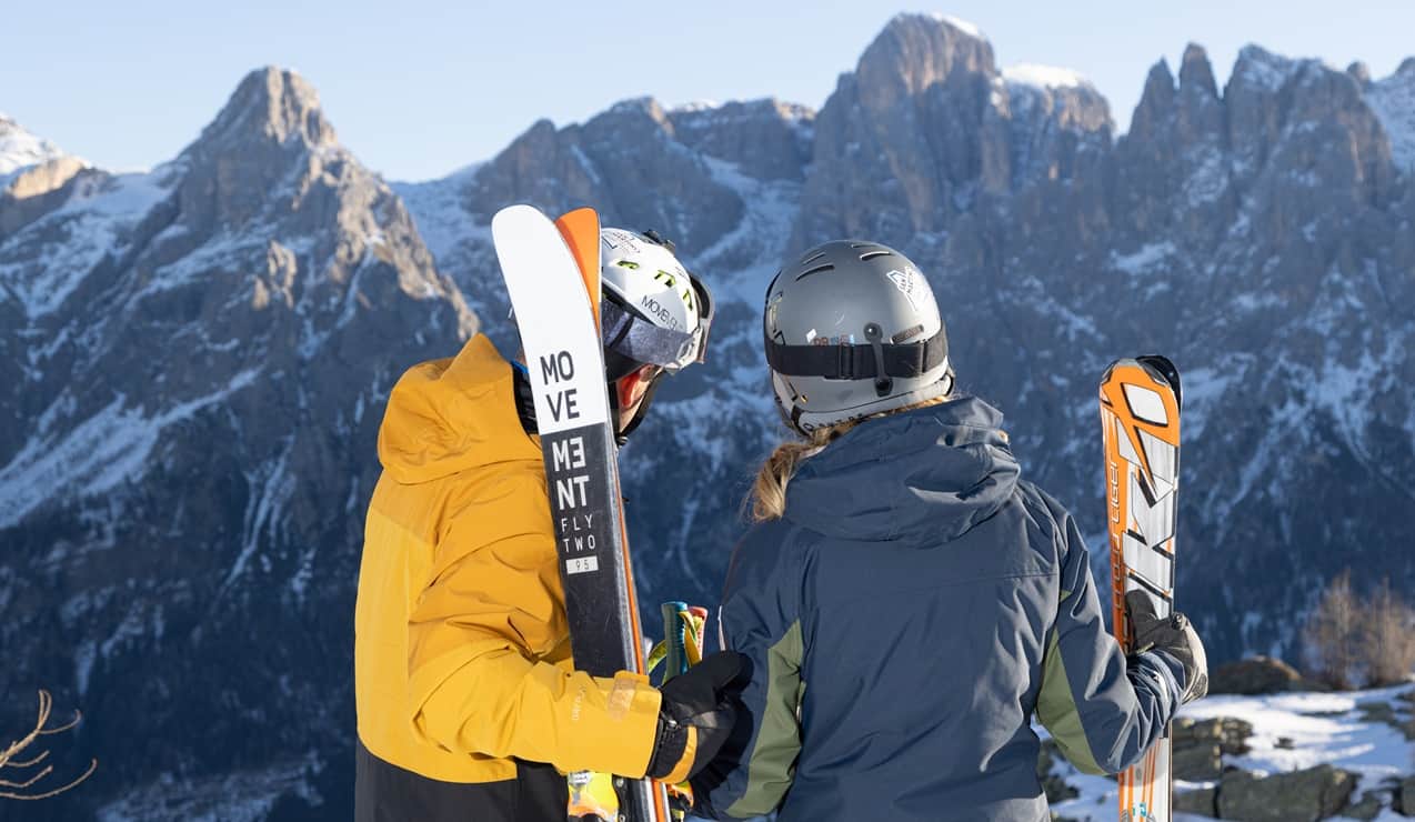 Due sciatori con casco e giacche che tengono gli sci sulle spalle, osservano le montagne innevate