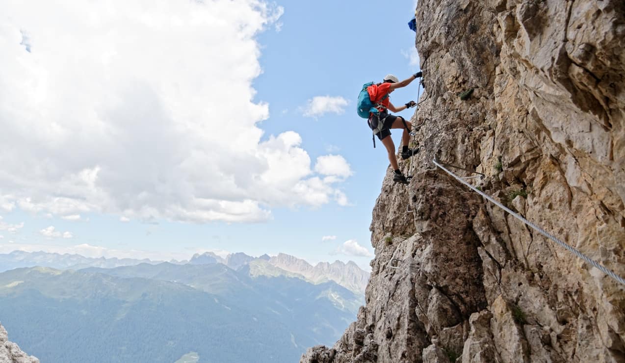 Alpinista con casco e zaino che scala una parete rocciosa attaccato a una via ferrata, montagne e cielo sullo sfondo.