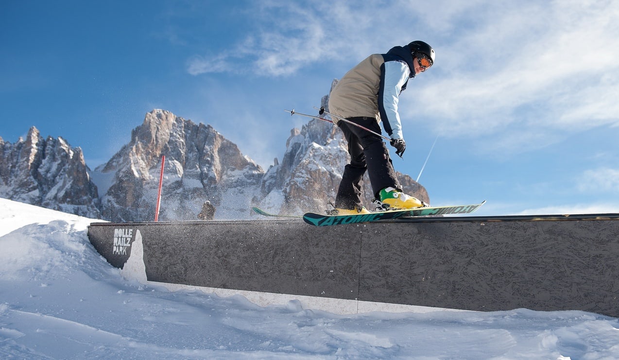 Sciatore scivola su un rail dello snowpark, montagne rocciose innevate sullo sfondo e cielo azzurro