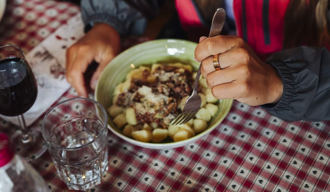 Mano con forchetta che mangia gnocchi con sugo in ciotola su tavola a quadretti, bicchieri vicini