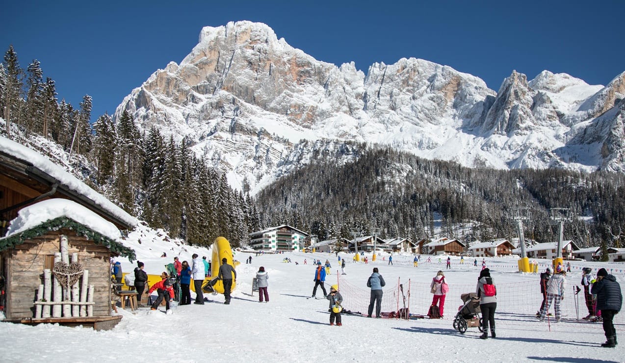 Persone in area innevata di comprensorio sciistico con chalet, alberi e grandi montagne innevate sullo sfondo