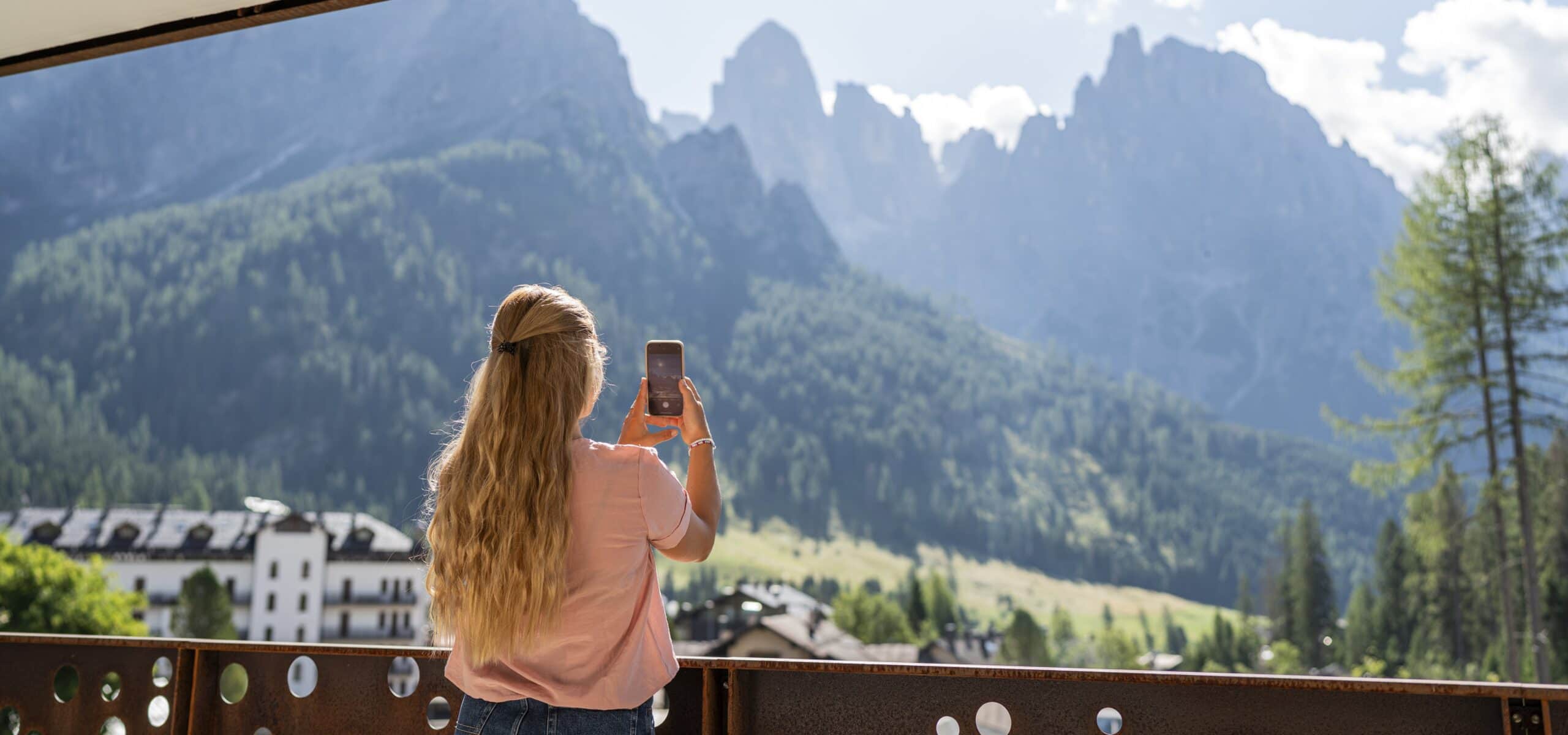 Donna di schiena scatta foto con smartphone dal balcone verso montagne e edifici alpini