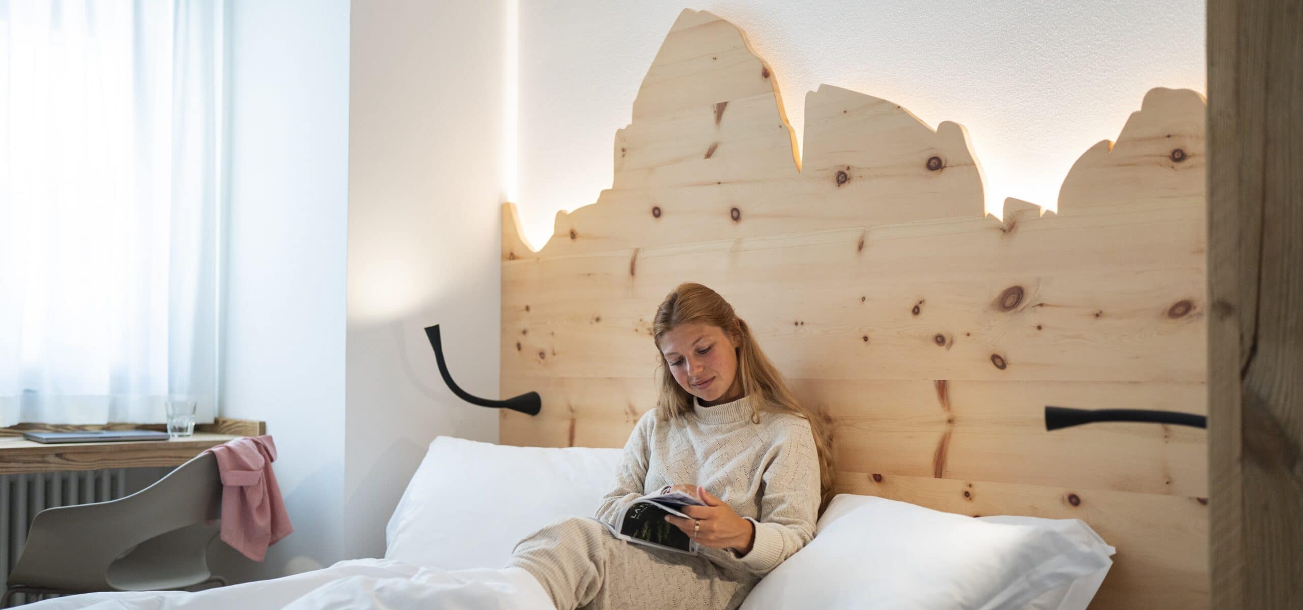 Donna seduta a letto che legge un libro, testiera in legno a profilo di montagna e finestra con tenda