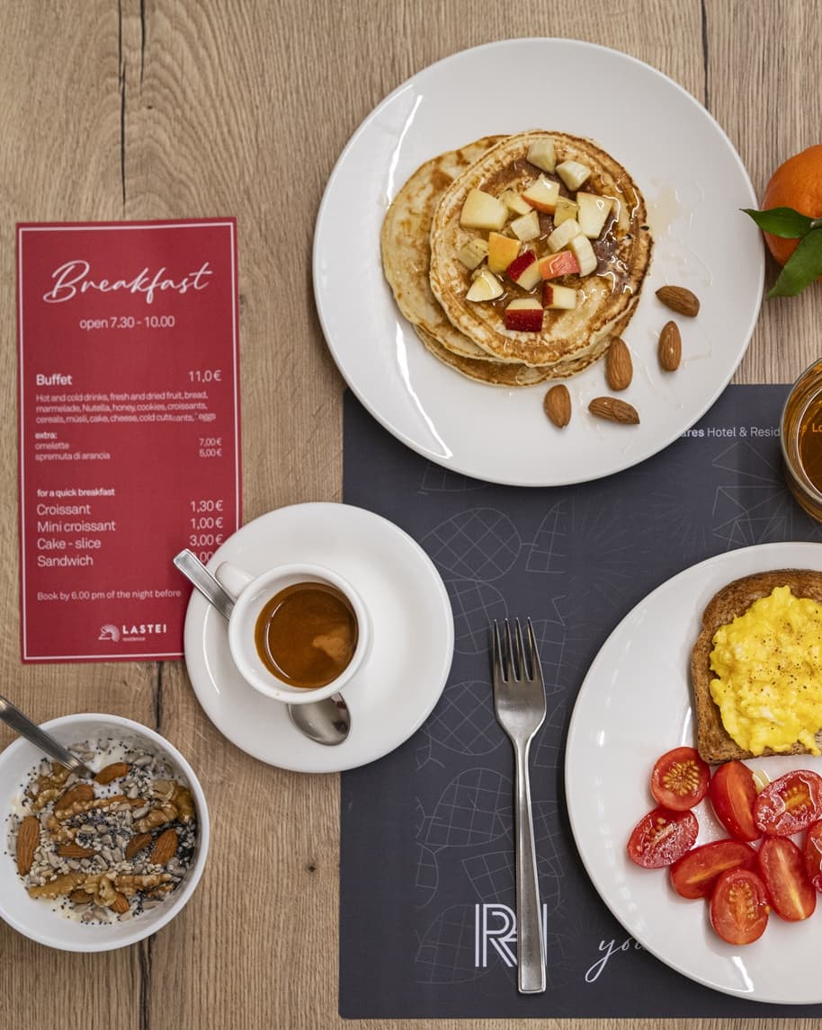 Pancake con frutta e mandorle su piatto, accanto espresso, uova strapazzate su pane, pomodorini e yogurt con noci