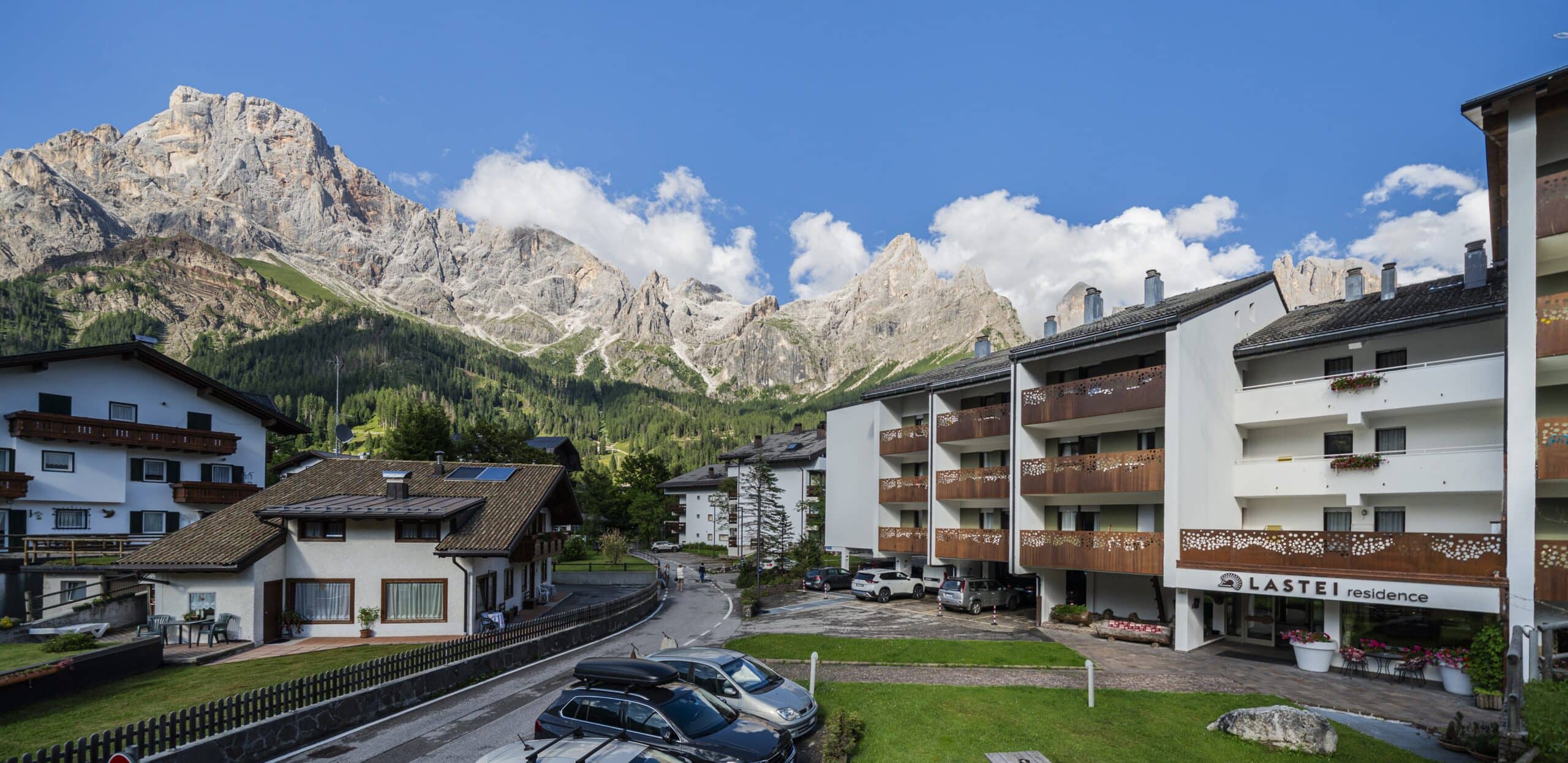 Edificio LASTEI Residence con balconi e parcheggio, piccoli edifici attorno e alte montagne rocciose sullo sfondo