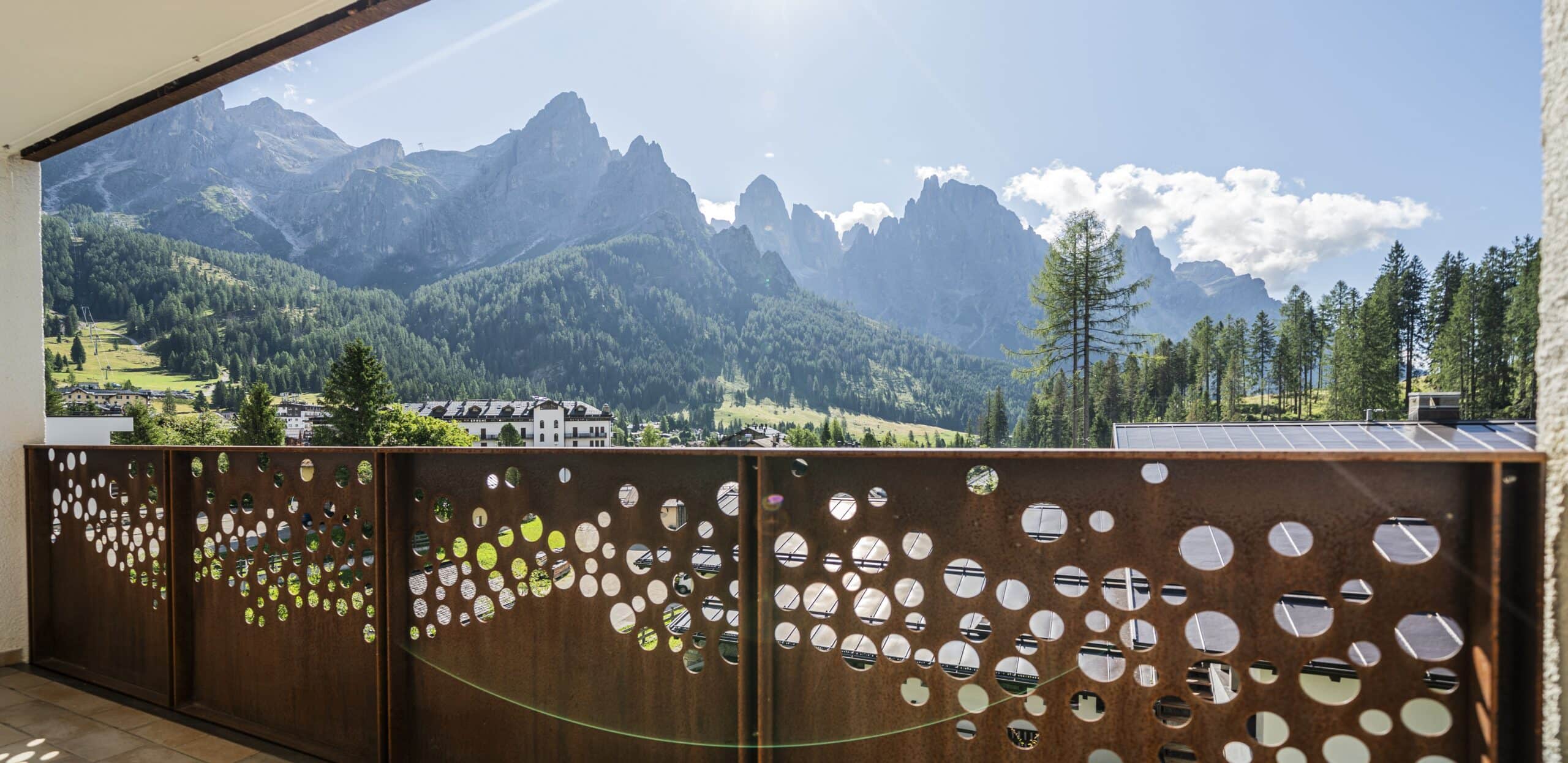Balcone con parapetto metallico traforato a fori circolari, davanti a bosco, edifici e montagne rocciose.