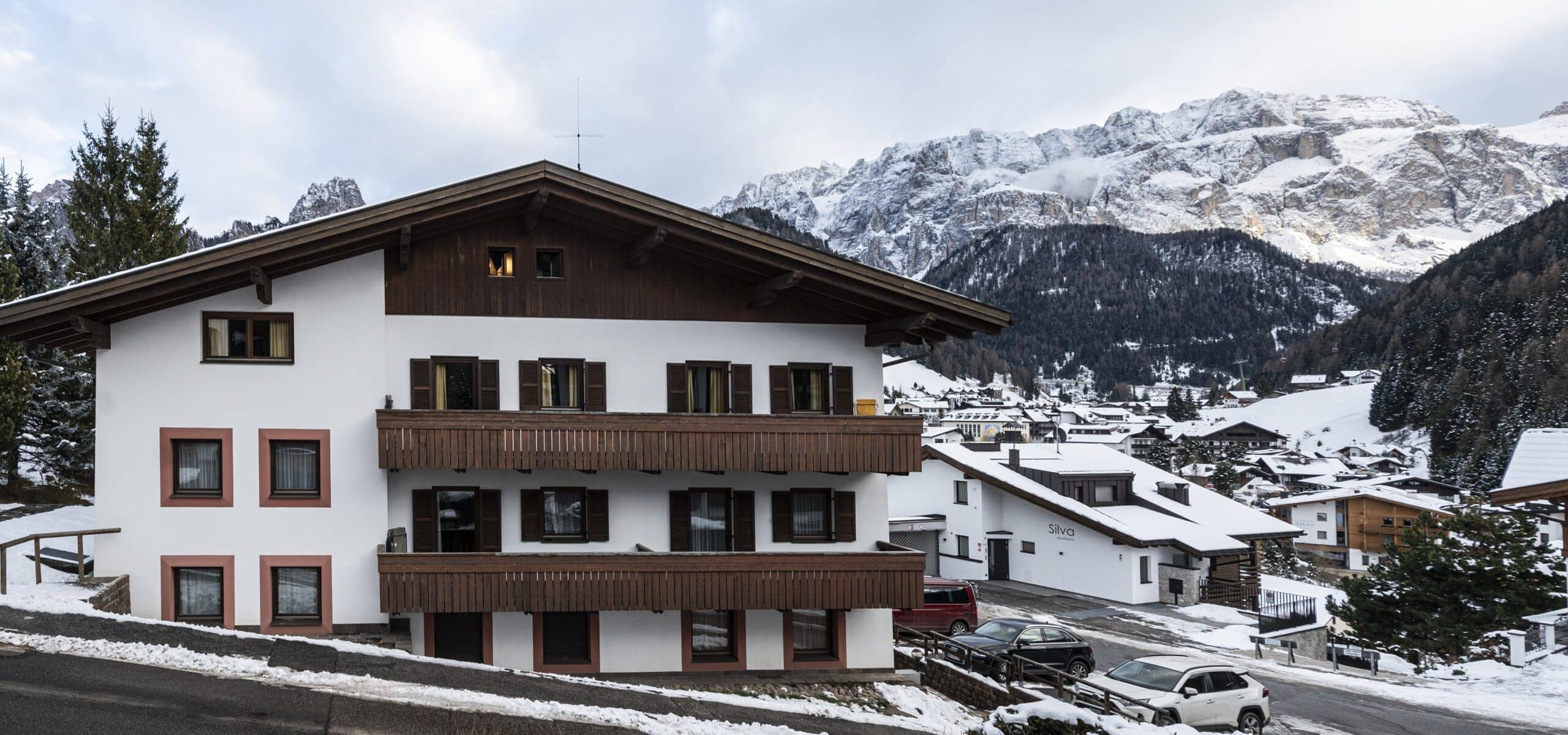 Chalet bianco con balconi in legno in paese innevato, auto parcheggiate e montagne rocciose sullo sfondo