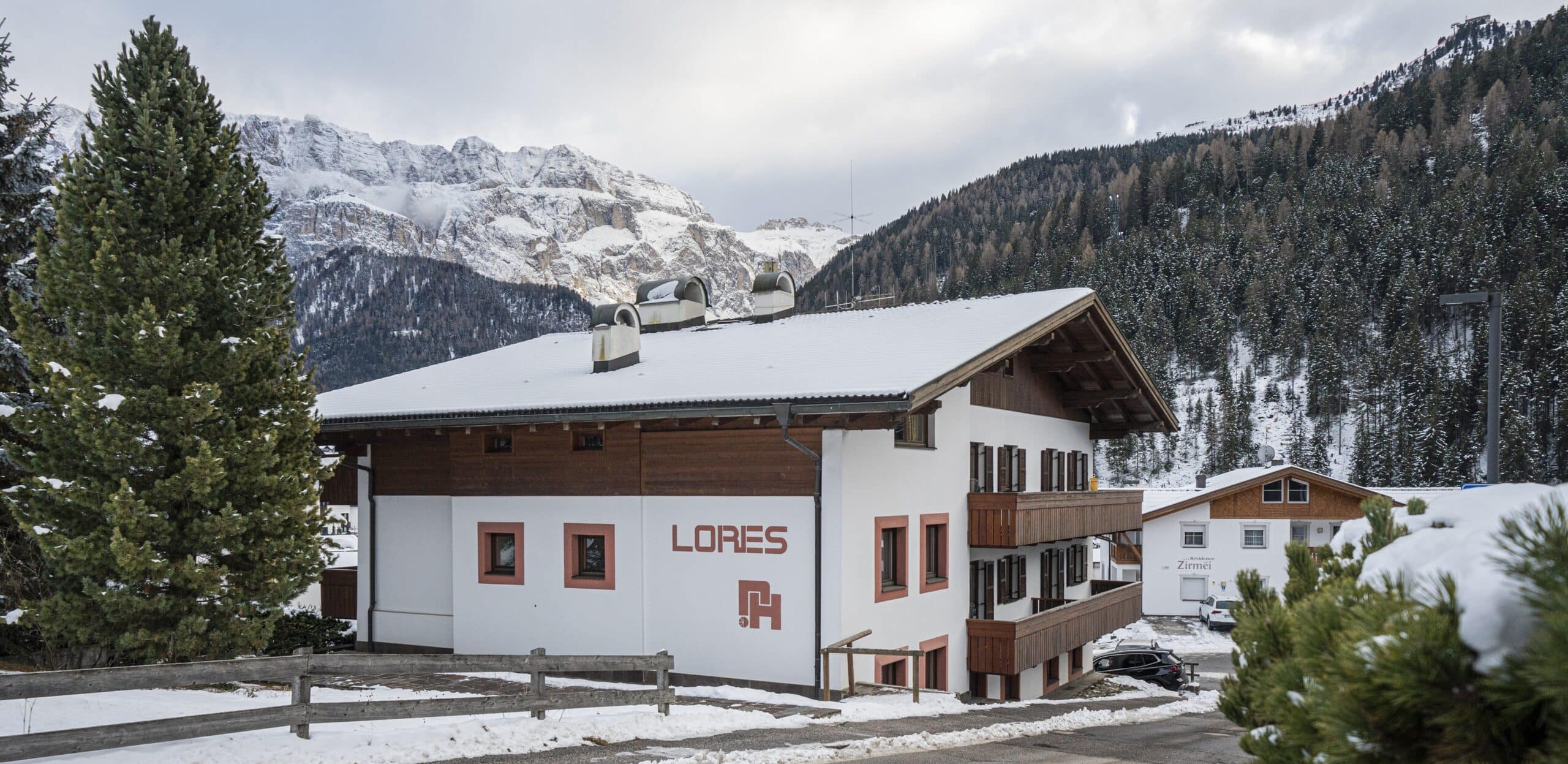 Edificio LORES con tetto innevato, balconi in legno, pini e montagne innevate sullo sfondo