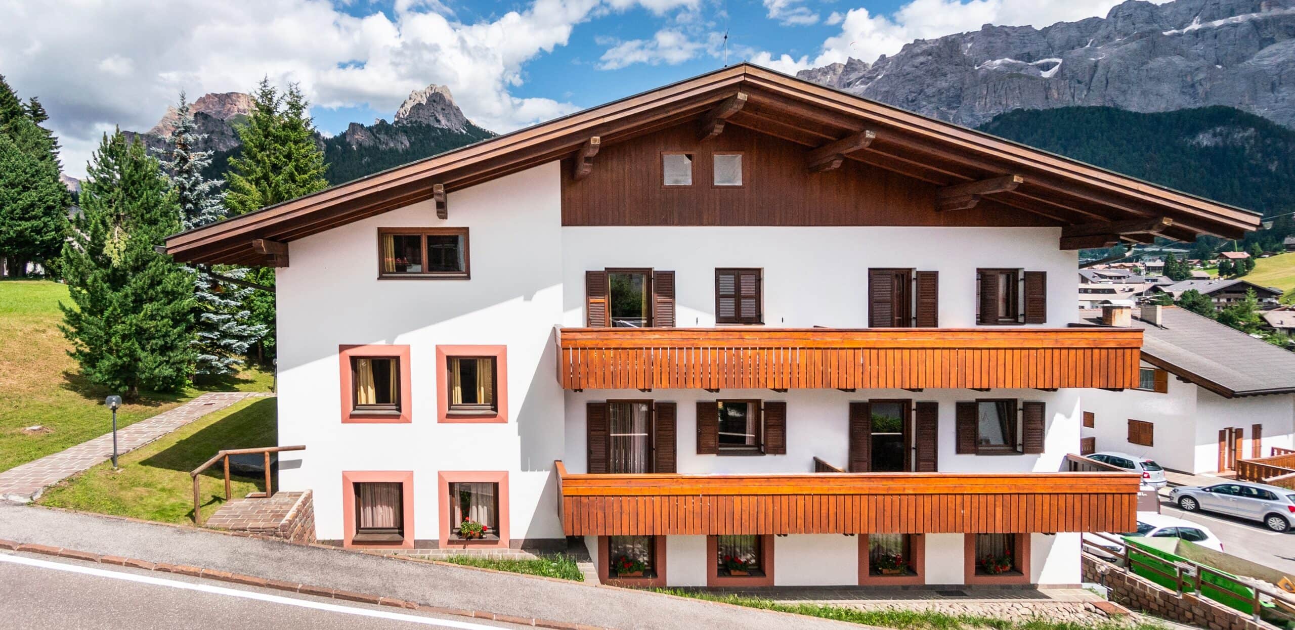 Edificio stile chalet bianco con balconi e persiane in legno, prato e montagne rocciose sullo sfondo.