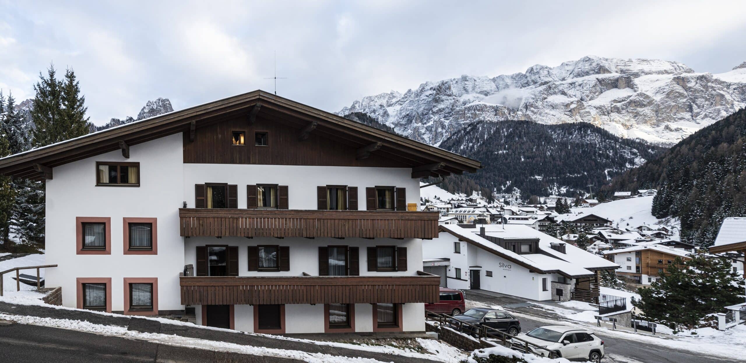Chalet a tre piani con balconi in legno su strada innevata, borgo e montagne innevate sullo sfondo