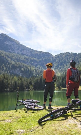 Due ciclisti con casco e zaino fermi sulla riva di un lago alpino, biciclette sull'erba e montagne boschive