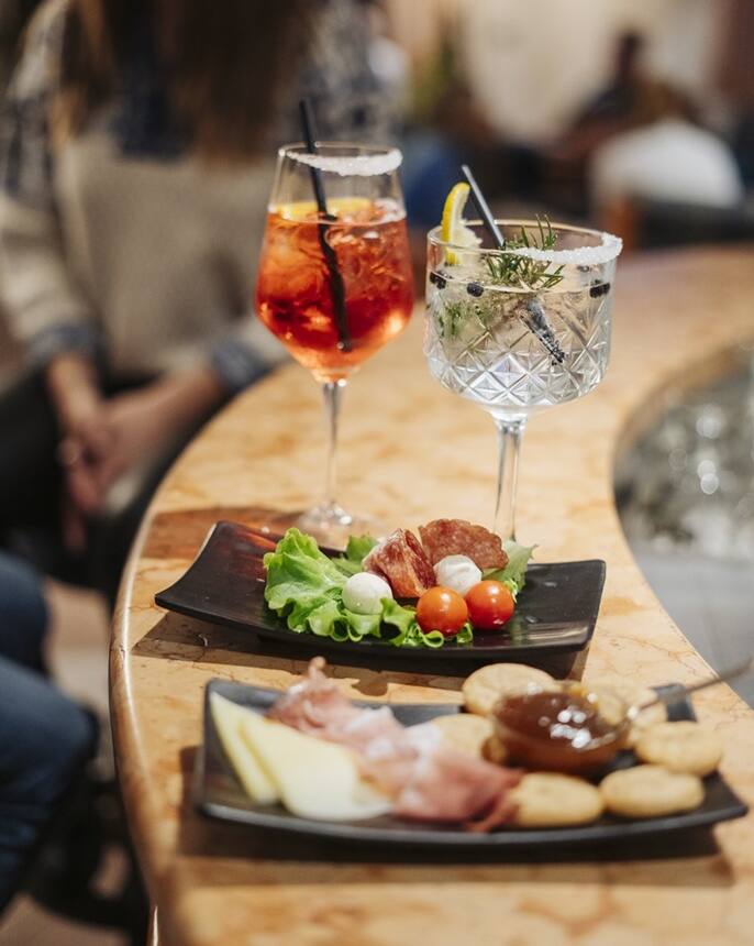 Zwei Cocktails auf Bartresen, davor Teller mit Salami, Mozzarella, Tomaten, Käse, Schinken und Crackern