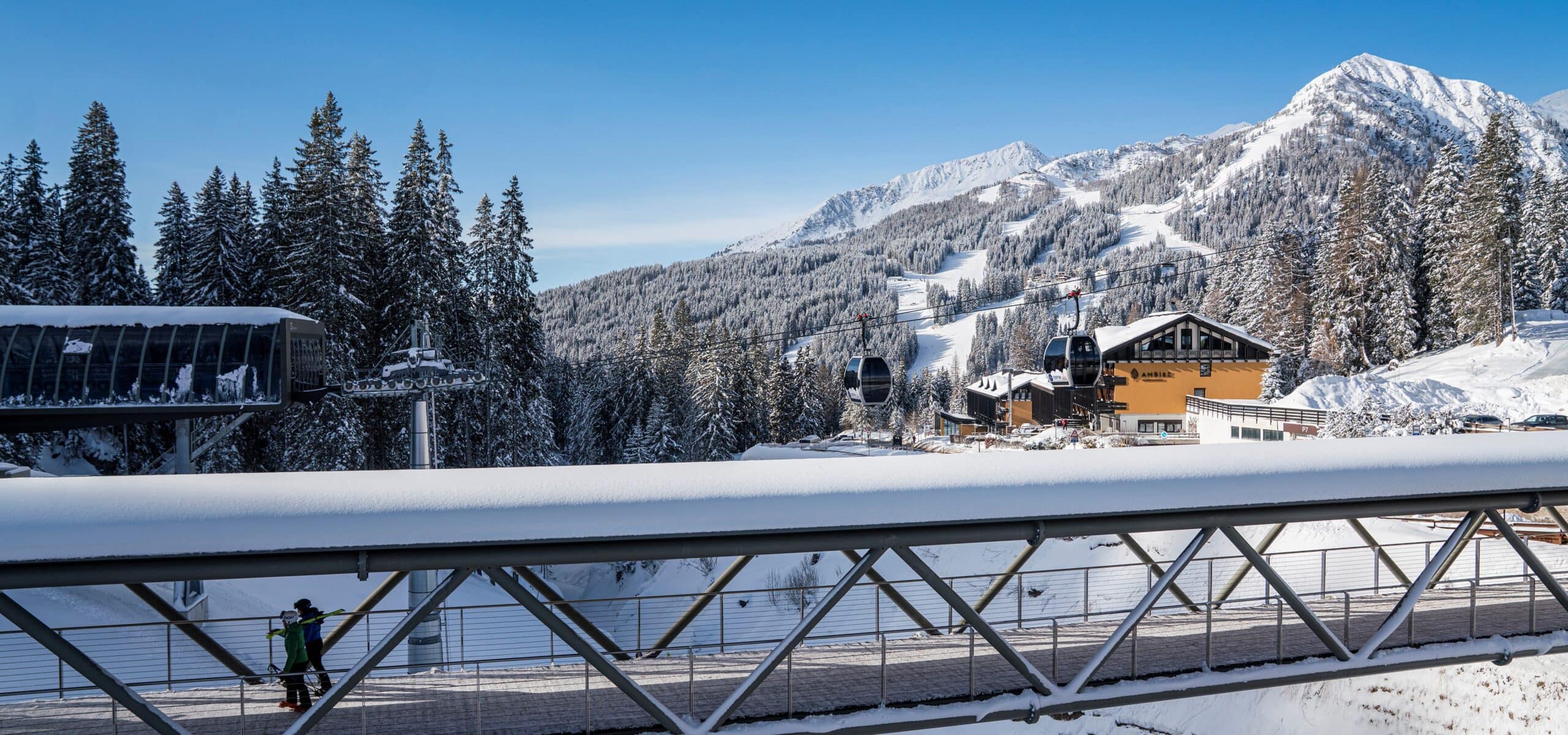 Cabine della funivia sopra una passerella innevata; due sciatori a piedi, pini e piste sulle montagne