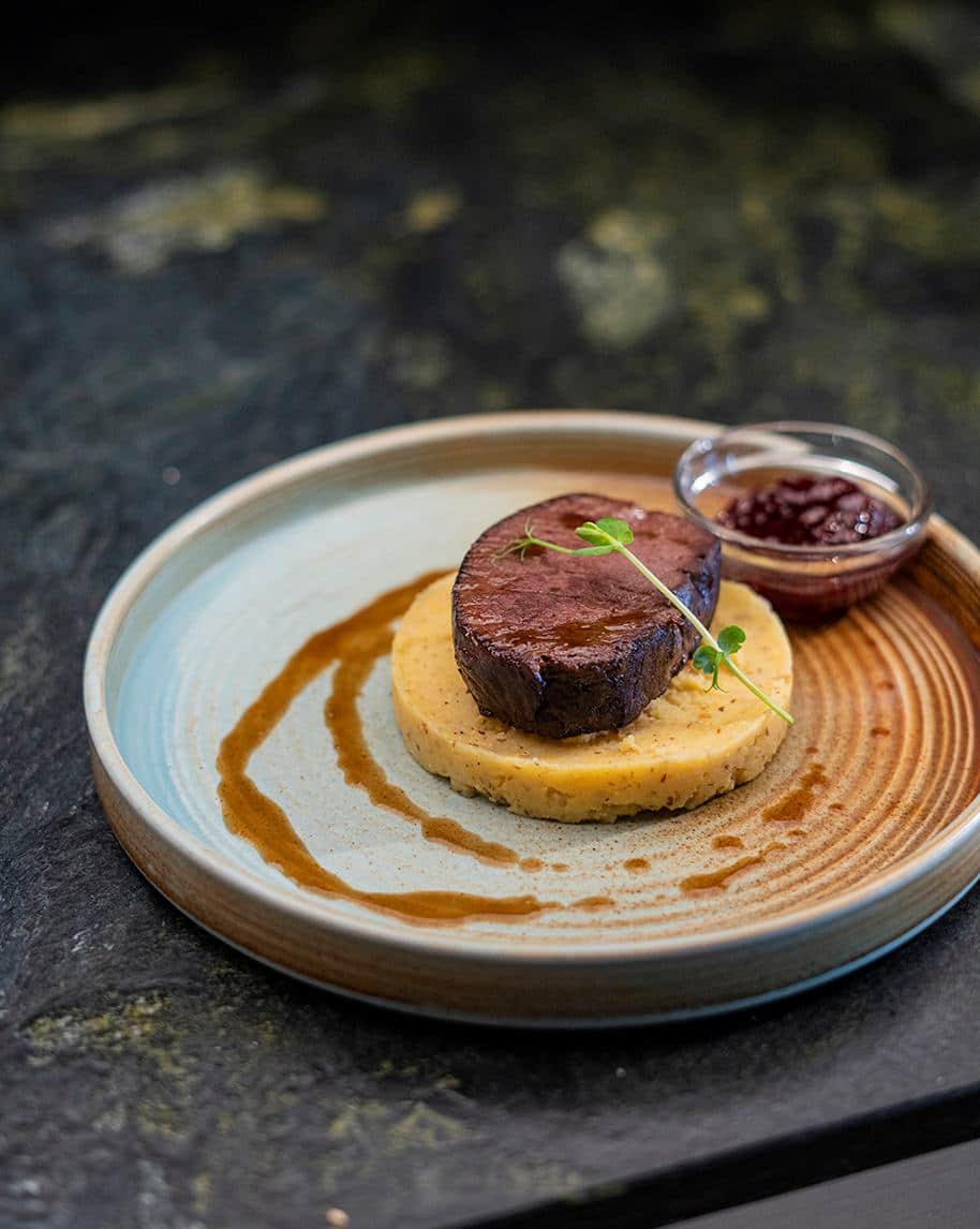 Filetto di carne scottato su disco di polenta con salsa a spirale e coppetta di composta su piatto in ceramica