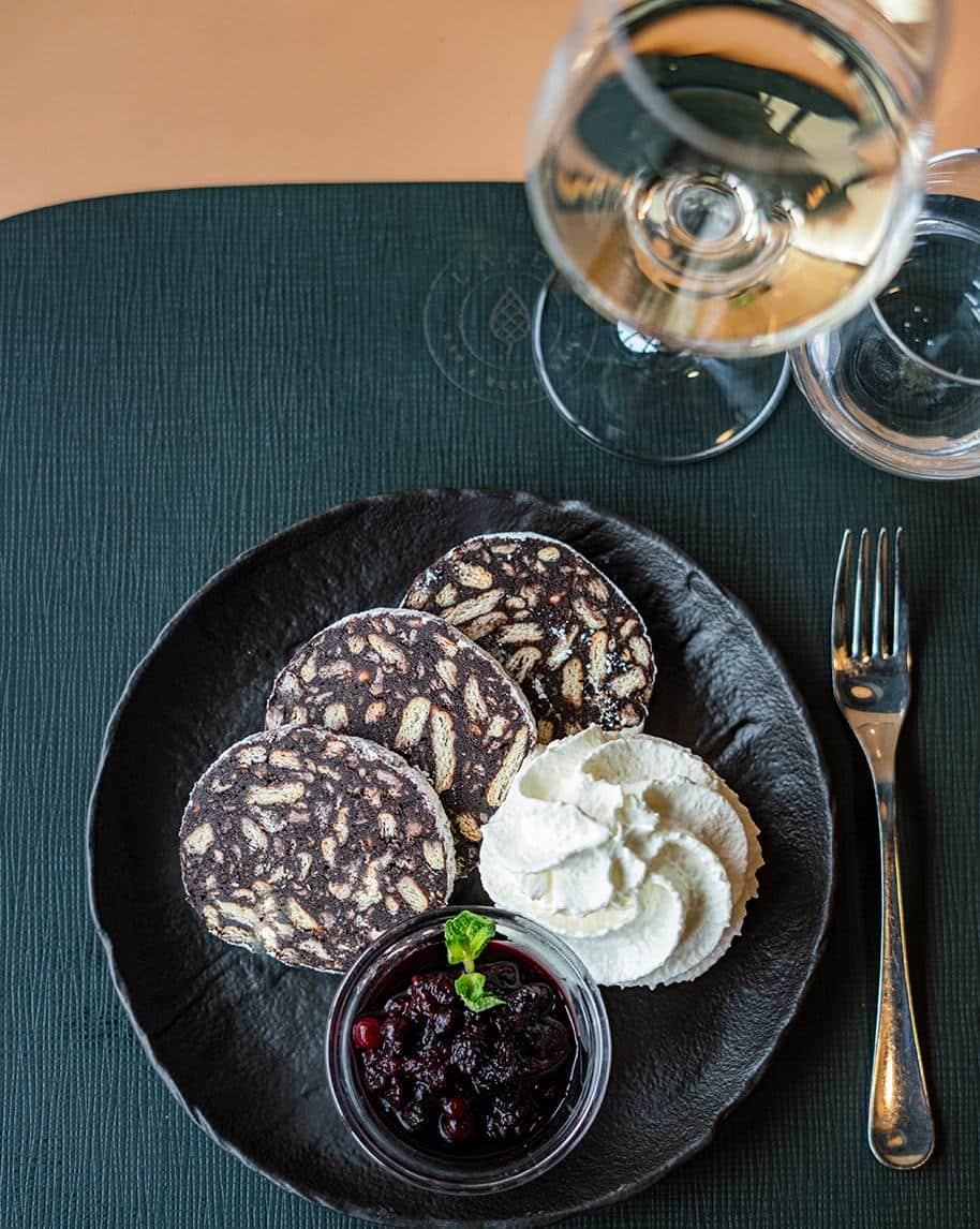 Tre fette di salame dolce al cioccolato, panna montata e composta di frutti rossi su piatto nero, bicchiere di vino
