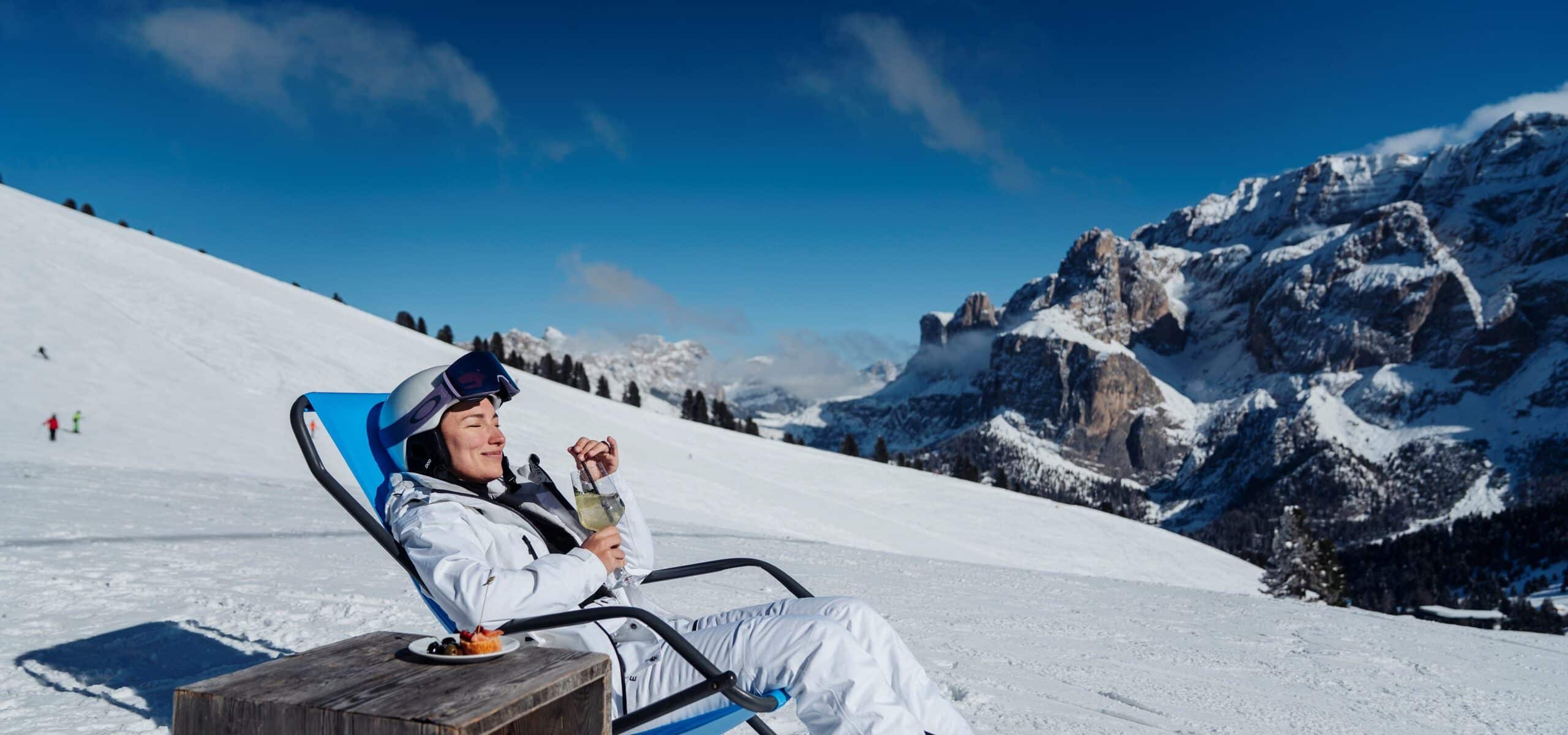 Persona in tuta da sci su sdraio blu su pendio innevato che sorseggia vino, montagne innevate sullo sfondo