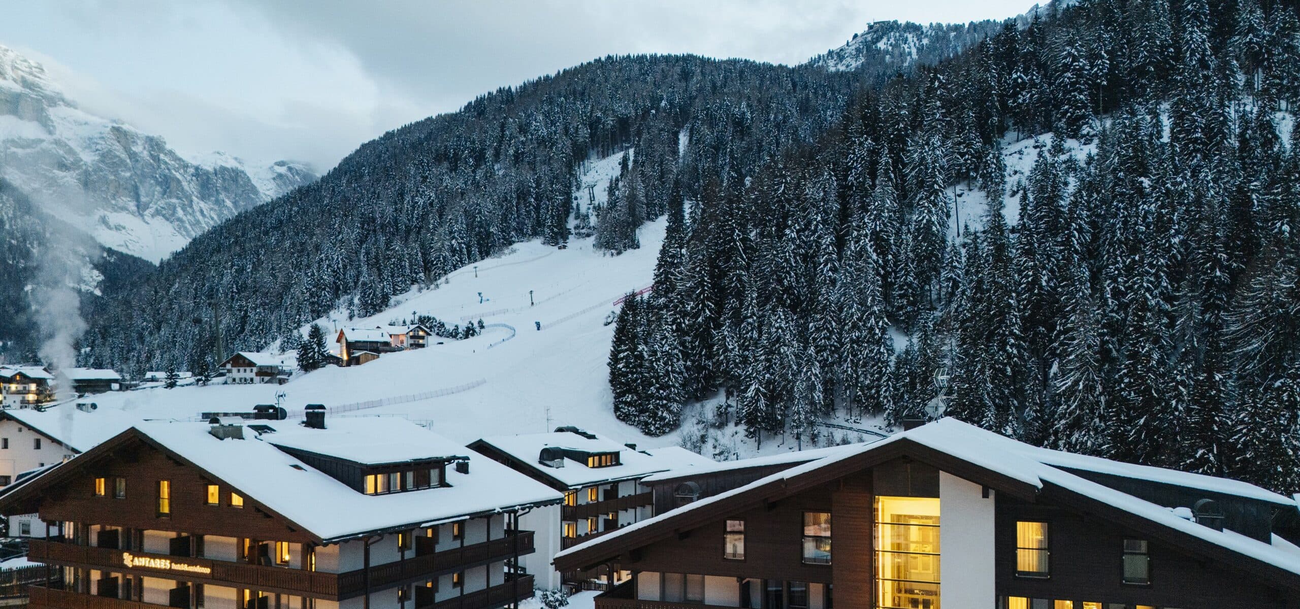 Chalet innevati con luci accese ai piedi di una pista da sci, sullo sfondo bosco e montagne