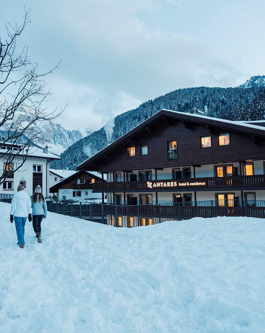 Edificio di Antares hotel & residence, due persone camminano nella neve con montagne innevate sullo sfondo