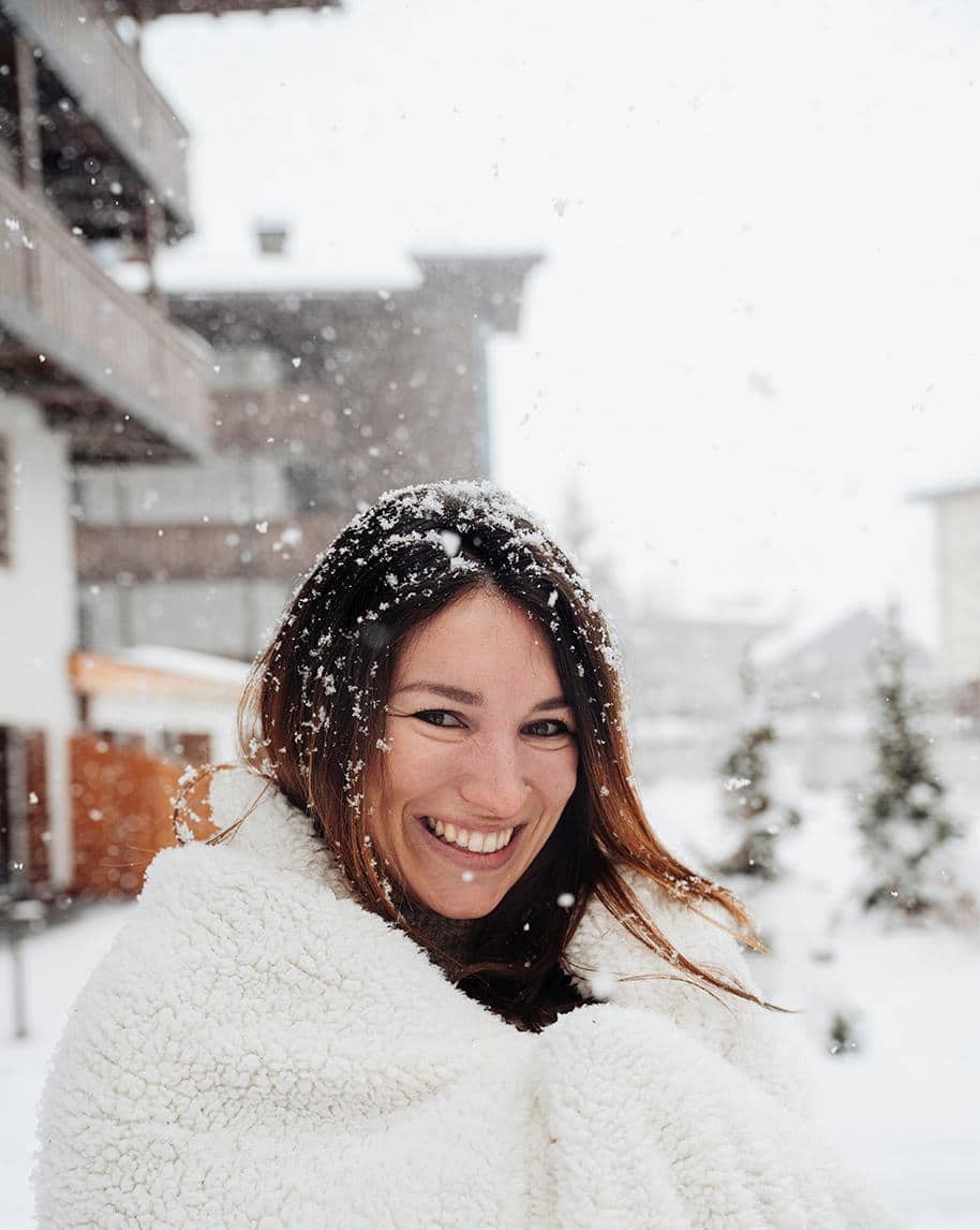 Donna sorridente avvolta in una coperta bianca, con fiocchi di neve tra edifici e alberi innevati