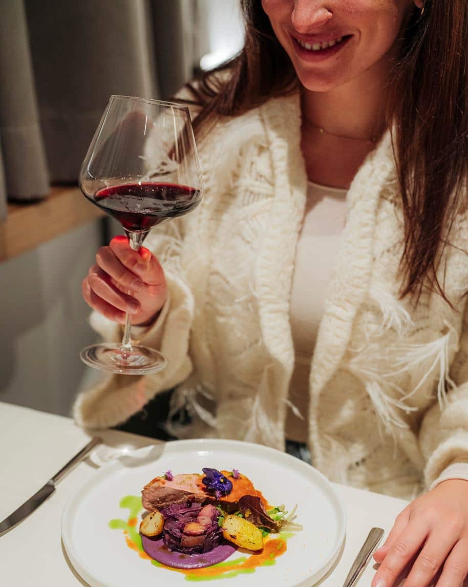 Donna sorride tenendo un bicchiere di vino rosso sopra un piatto gourmet su tavolo bianco